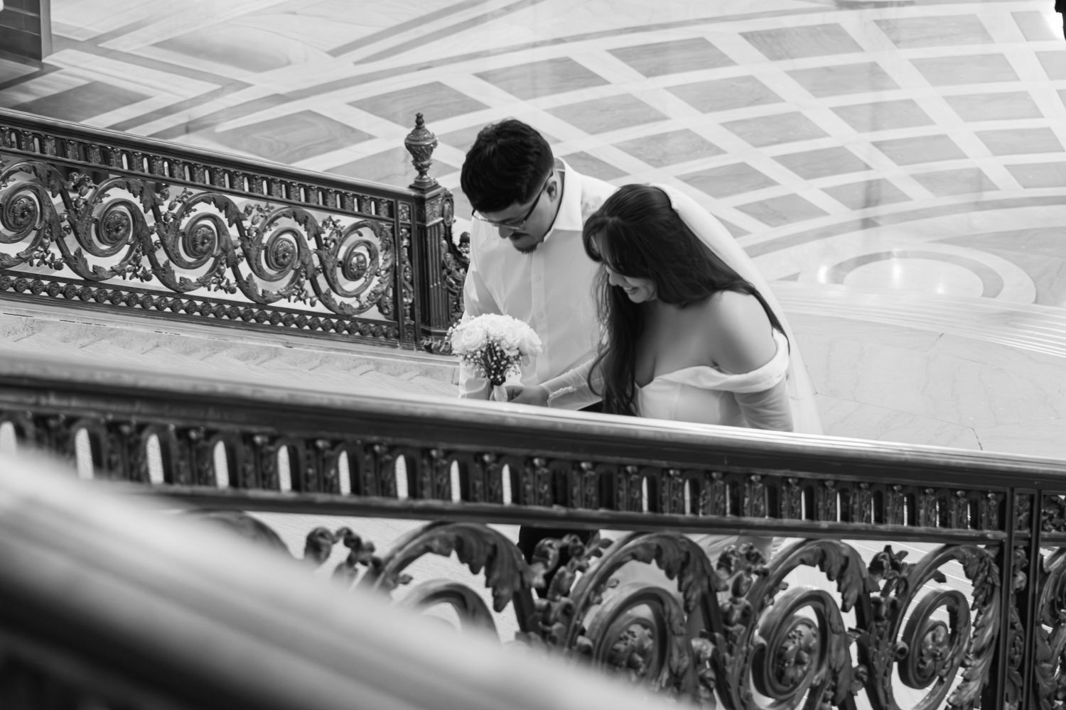 Black and white photo of a couple sharing a tender moment on an ornate staircase railing, bride holding a bouquet.
