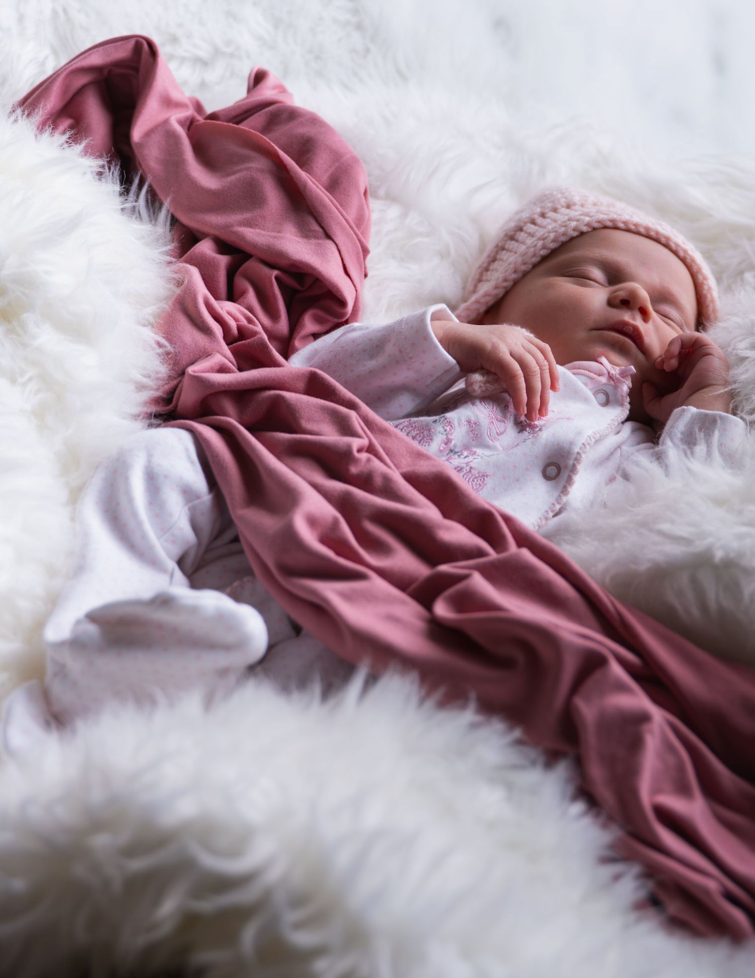 Newborn baby sleeping peacefully on white fluffy blanket wrapped in a soft pink knit hat and dusty rose swaddle.