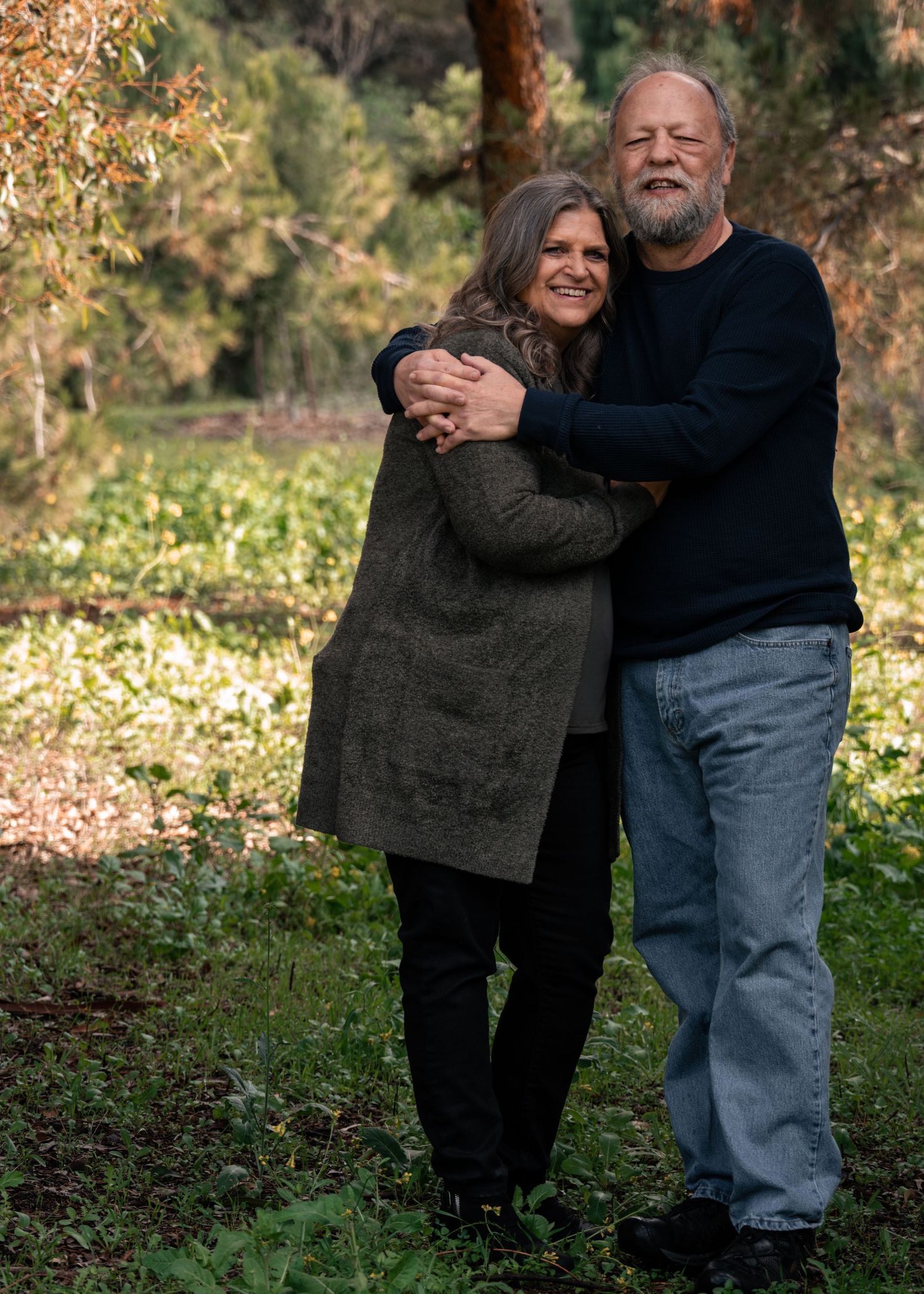 A mature couple embraces while standing outdoors in an autumn garden with fall foliage in the background.
