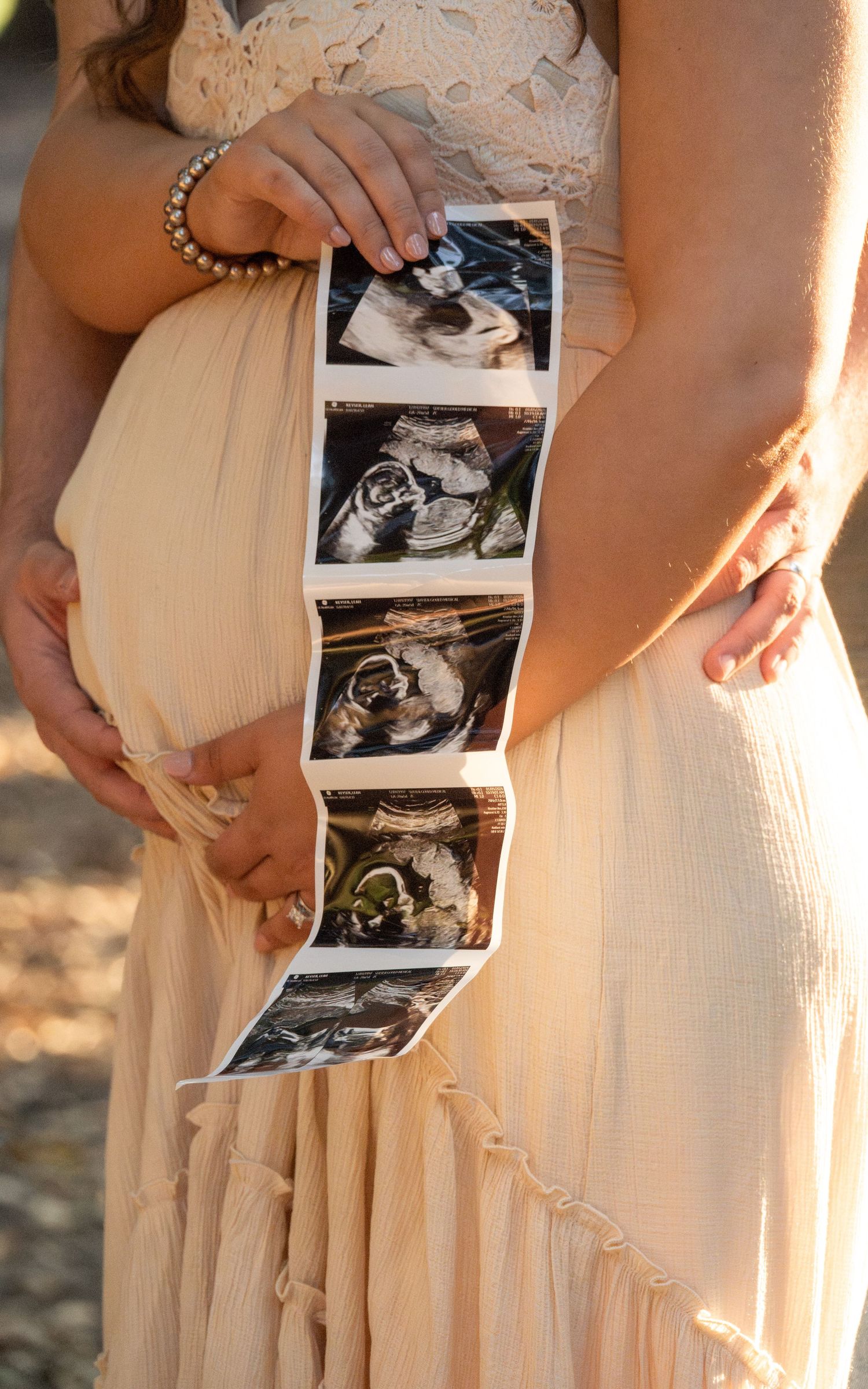 A person in a cream-colored dress holds ultrasound photos during a maternity photoshoot at sunset.
