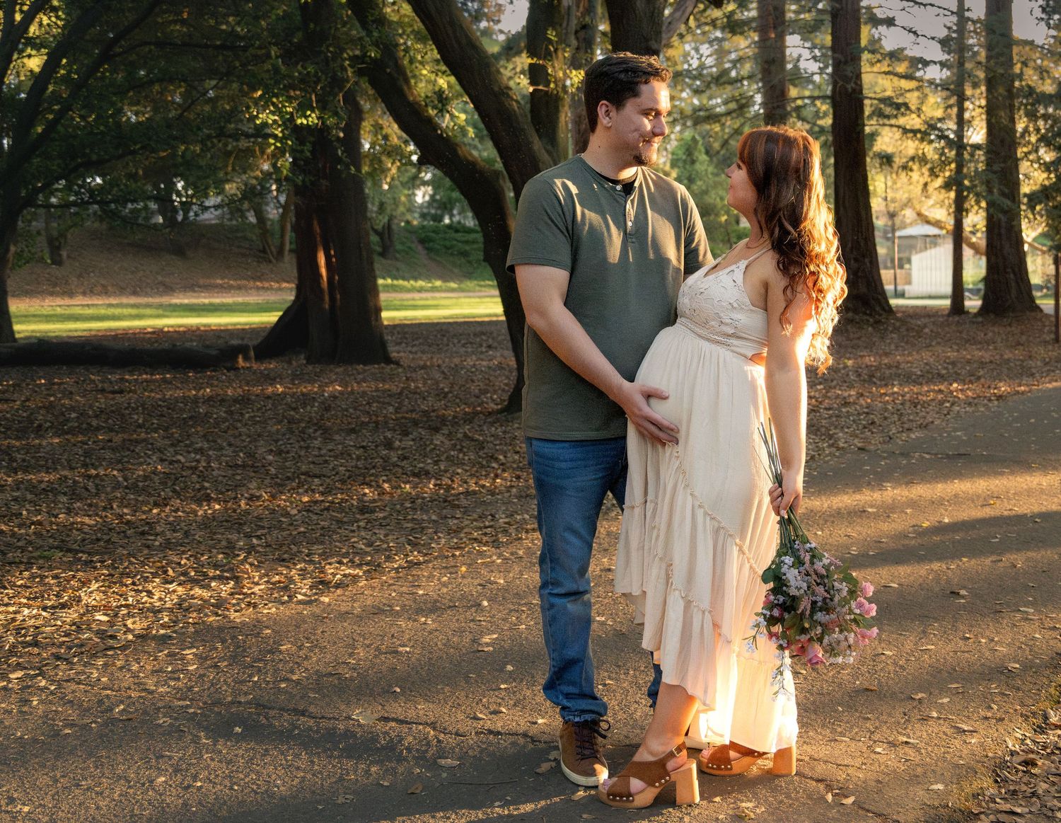 Pregnant couple sharing a tender moment in a sunlit park, woman in flowing dress holding flowers.