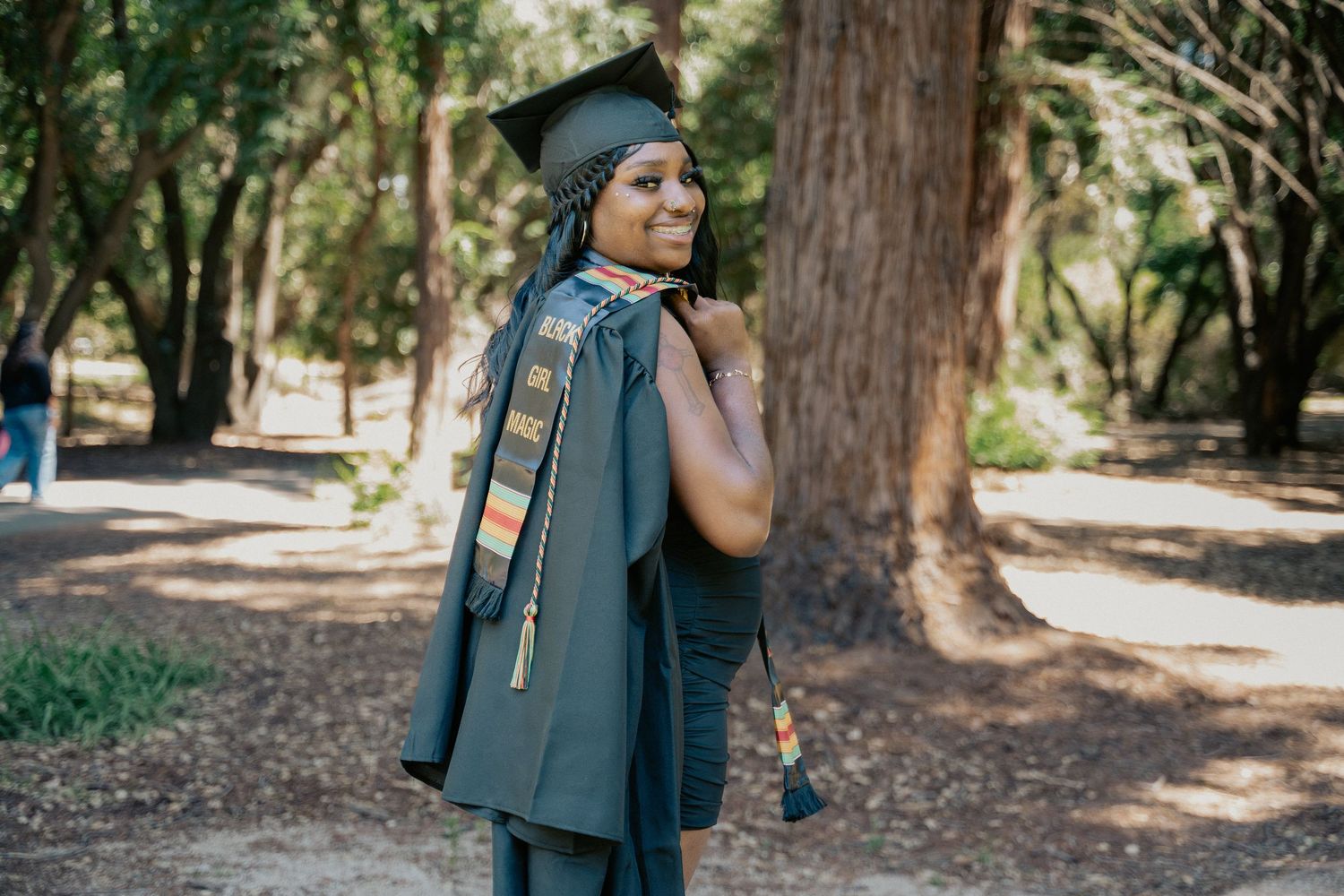 Smiling graduate in cap and gown poses outdoors among trees, wearing colorful stole and tassel.