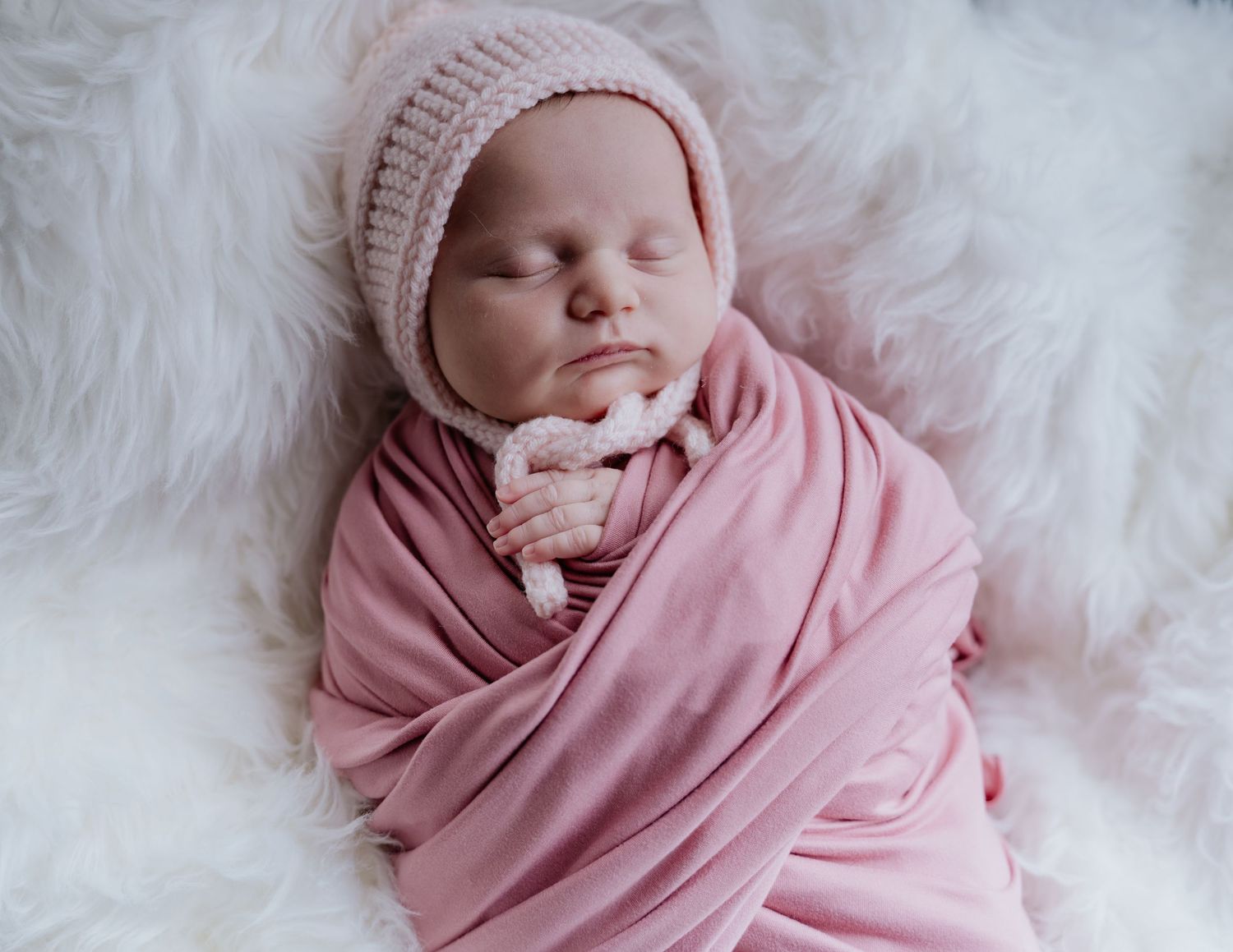 Newborn baby swaddled in pink wrap wearing knit bonnet sleeping peacefully on white fluffy blanket.