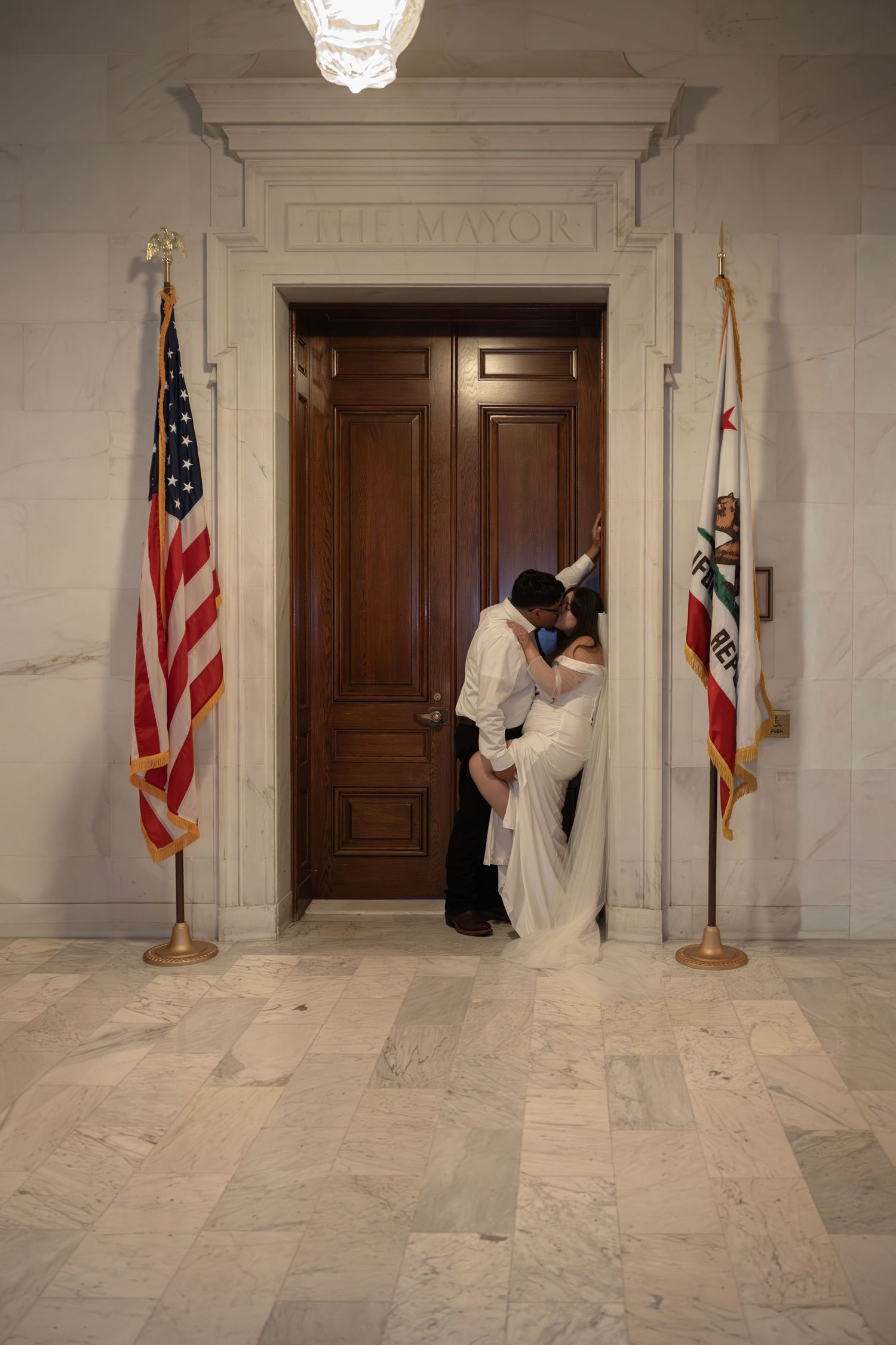Couple shares a kiss in front of The Mayor's office door flanked by American and California flags.
