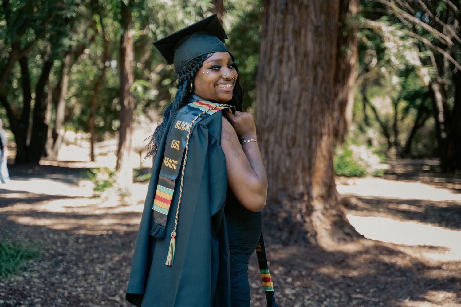 Smiling graduate in cap and gown with colorful stole poses outdoors among tall trees on graduation day.