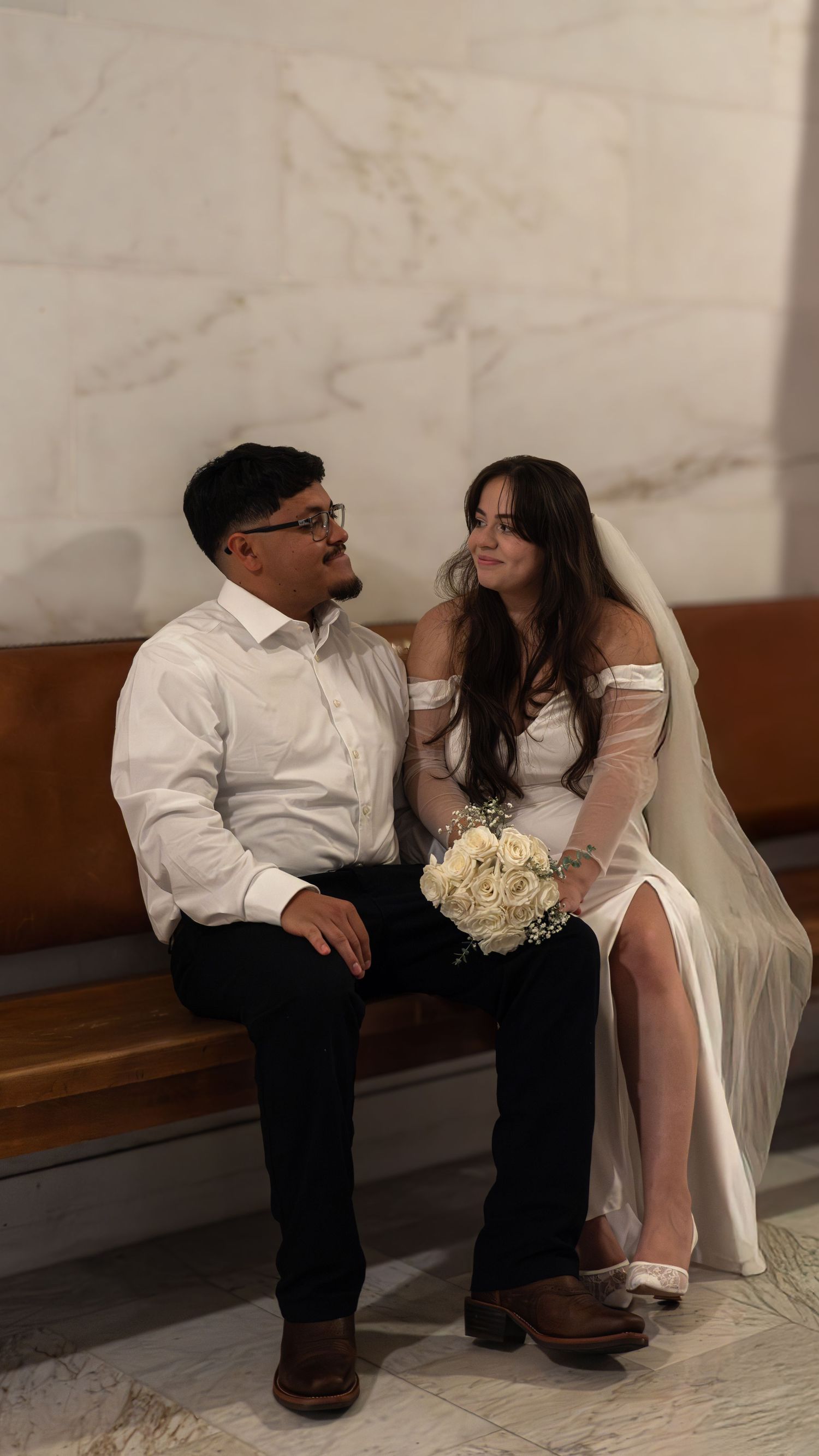 A couple in wedding attire sits on a bench, the bride holding white roses bouquet wearing a veil, groom in white shirt and black pants.
