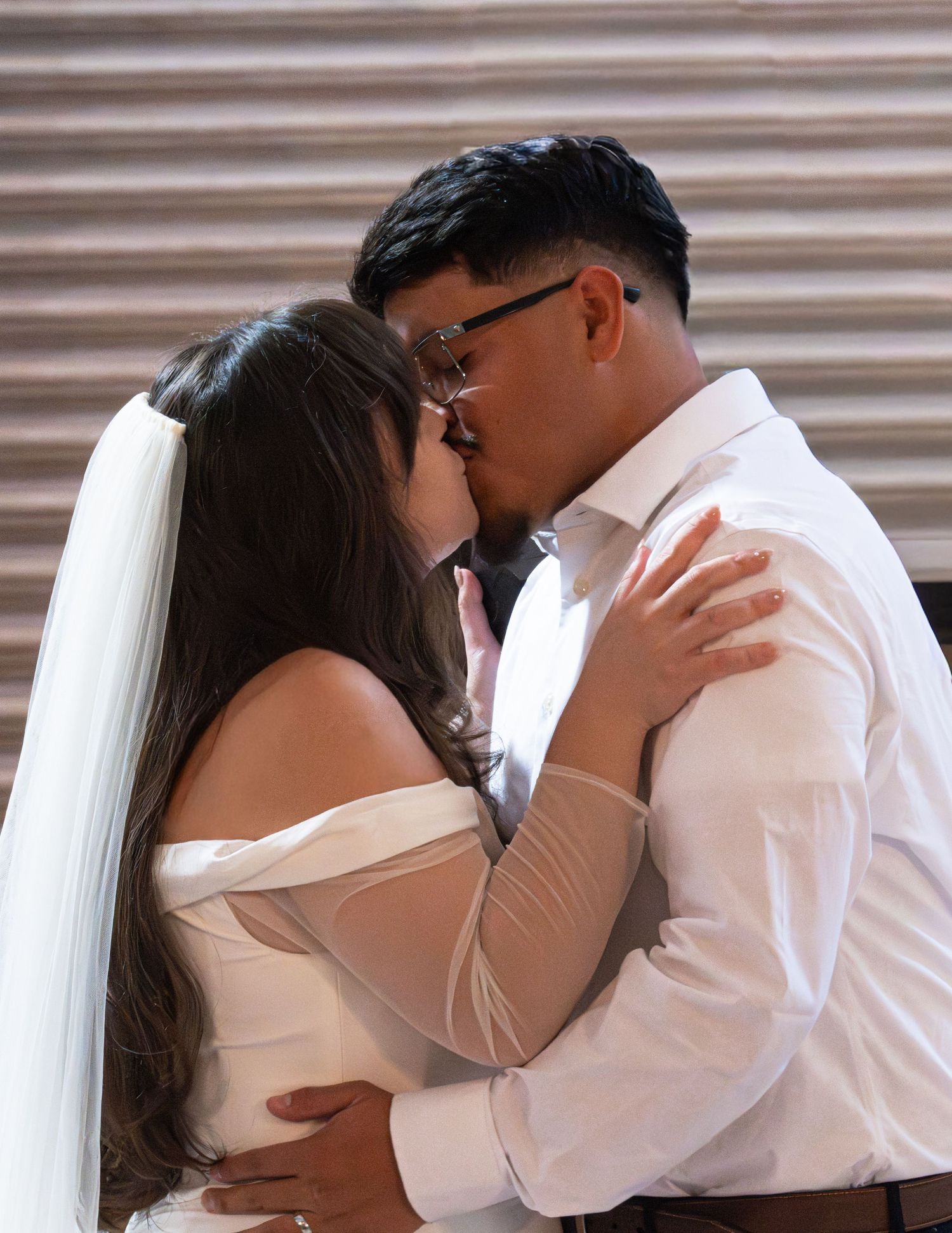 Bride and groom sharing their first kiss at wedding ceremony, bride wearing white off-shoulder gown and veil.