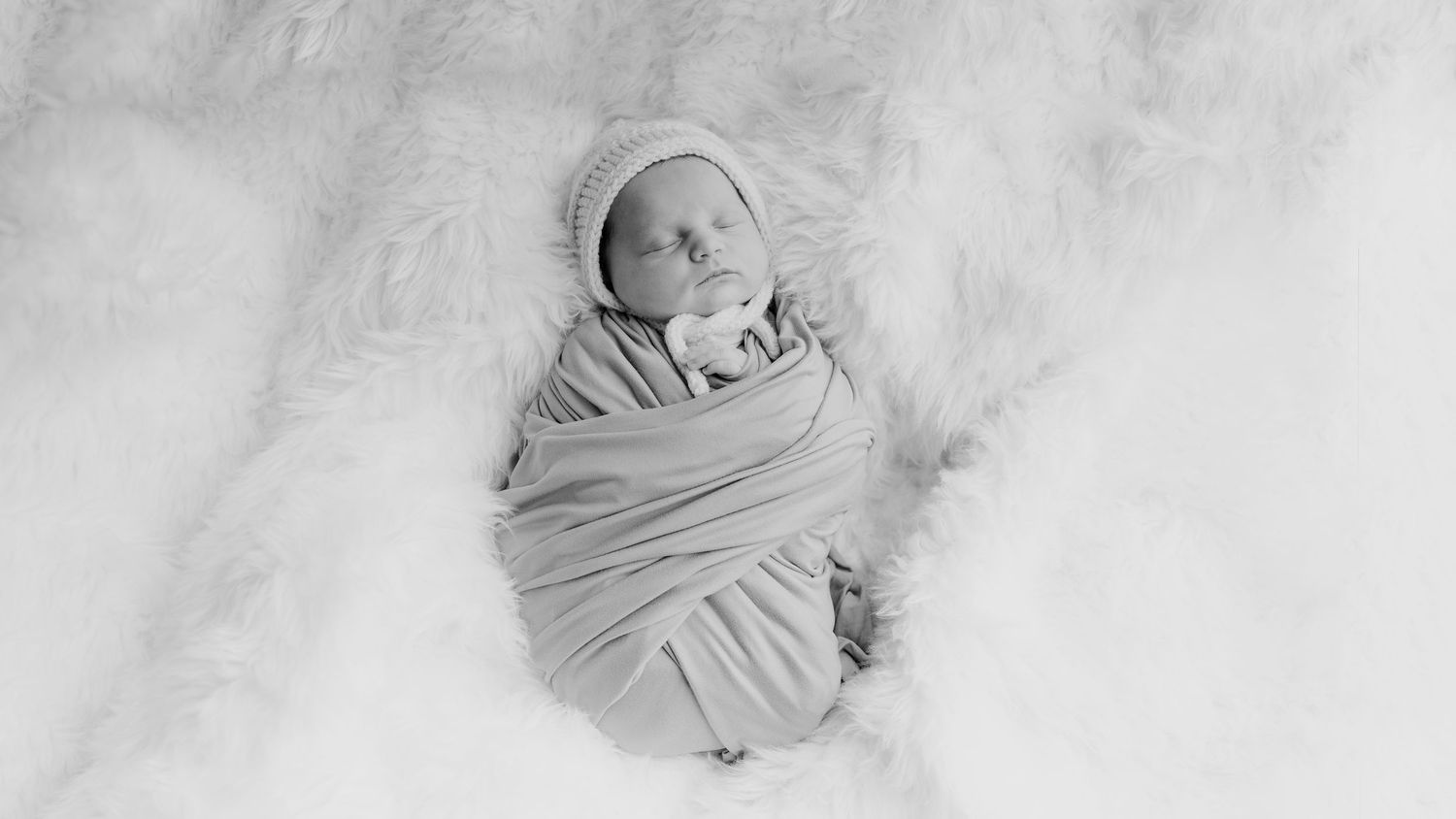 Black and white photo of a swaddled newborn baby wrapped in soft cloth lying on a fluffy white fur blanket in Newborn Photoshoot.