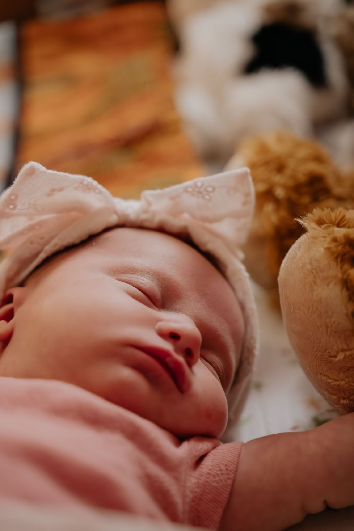 Sleeping newborn baby wearing a pink bow headband and outfit, lying next to a stuffed teddy bear.