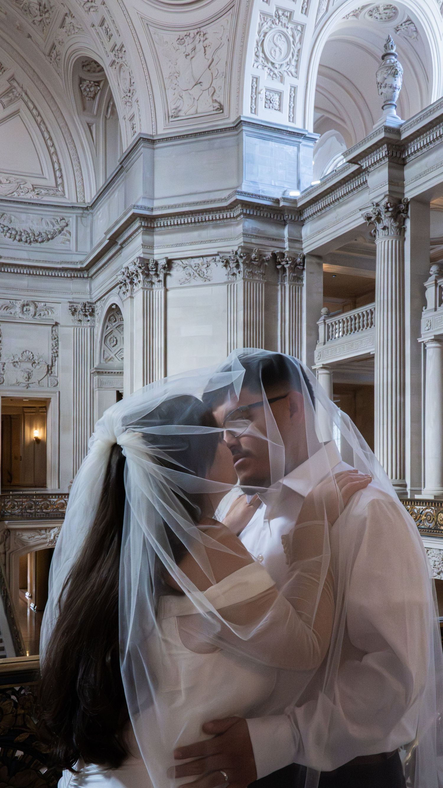 Bride and groom sharing a romantic kiss under a wedding veil in an elegant neoclassical hall.