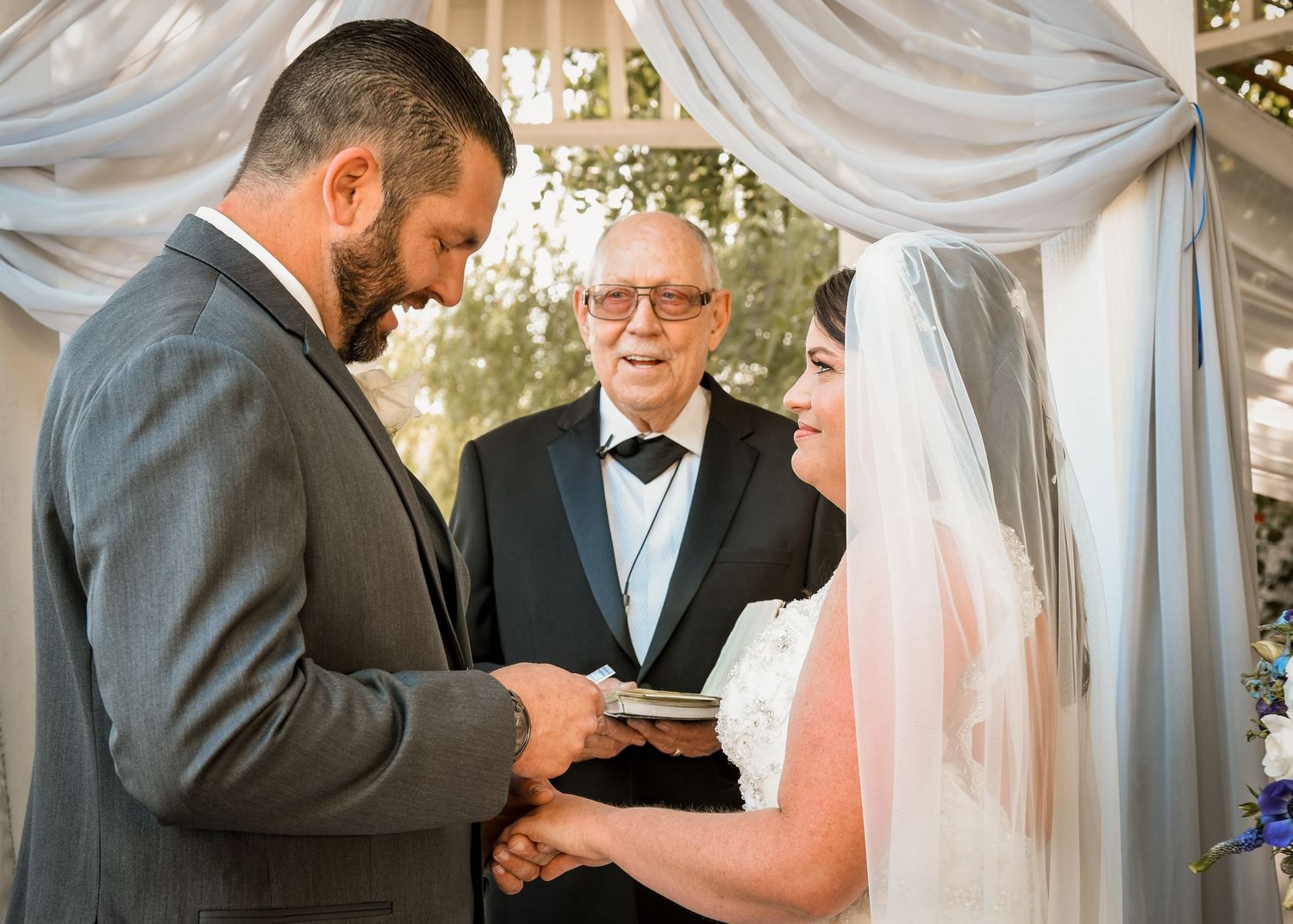 Groom places ring on bride’s finger during wedding ceremony under draped arch, with officiant smiling behind them. Emotional vow exchange.
