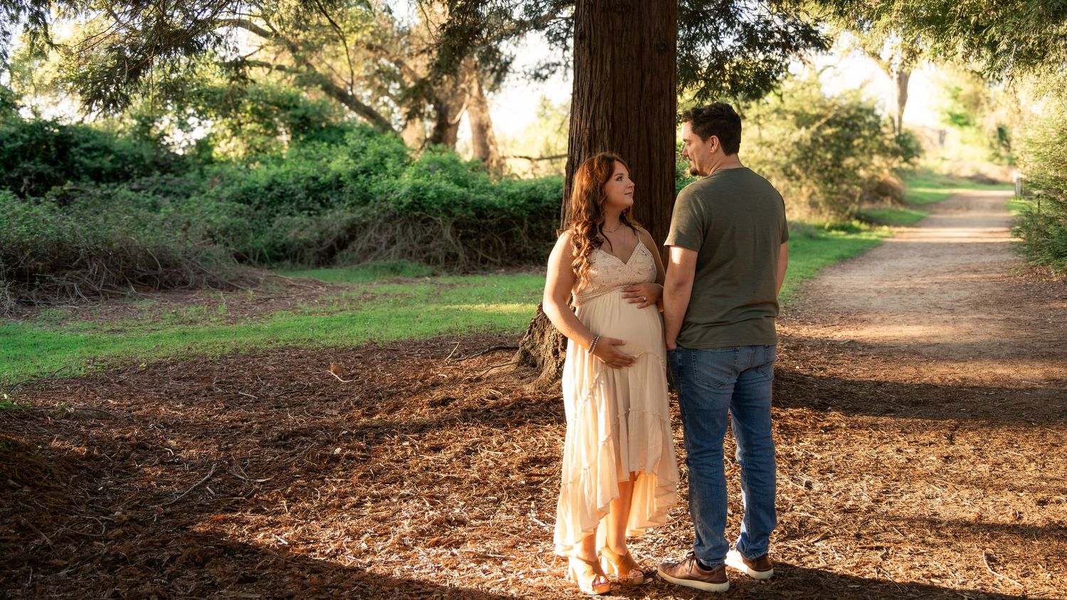 Pregnant couple sharing a tender moment under a large tree on a sunlit park path surrounded by lush greenery. Rocklin, CA