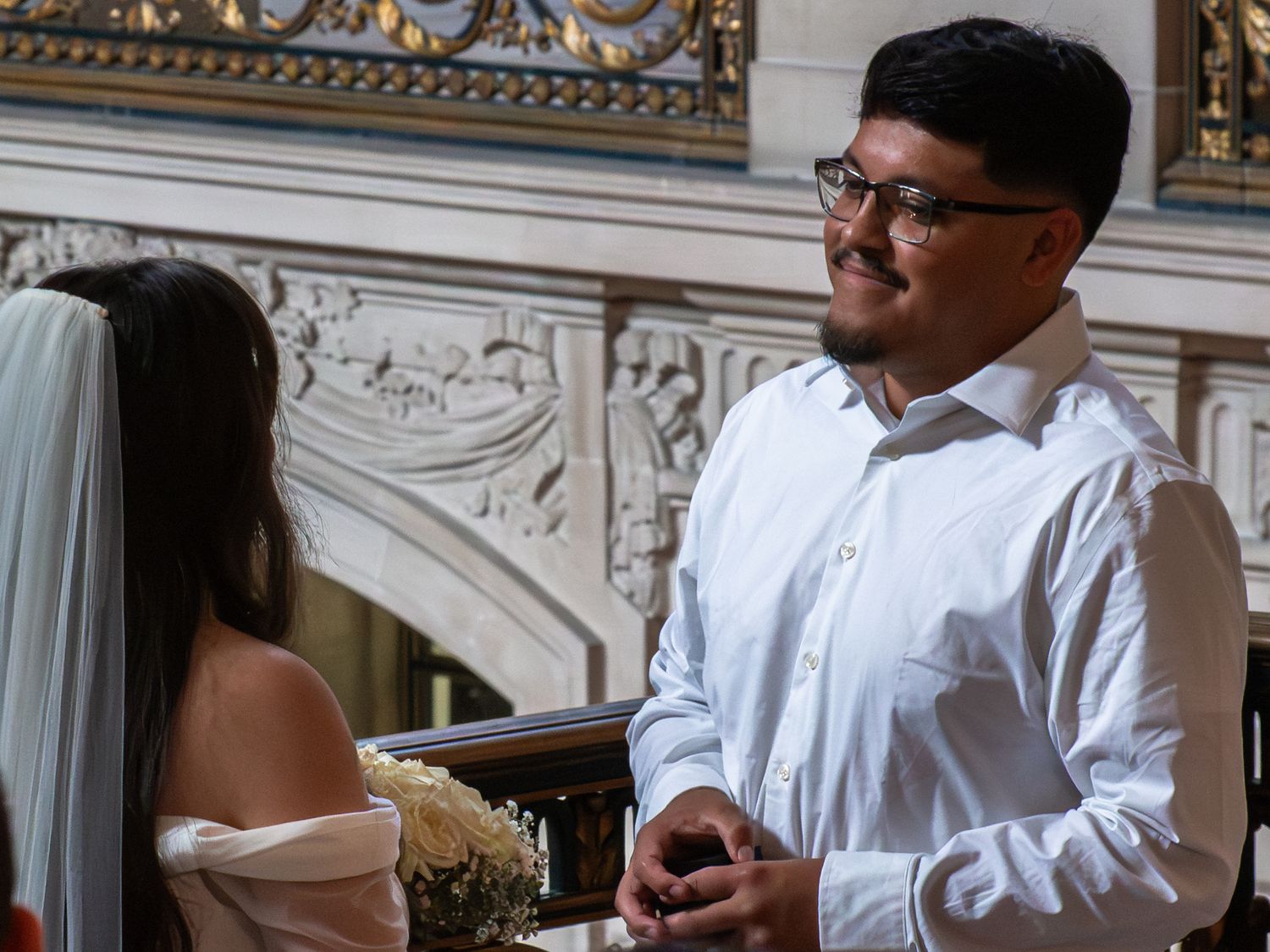 Groom in white shirt smiling at bride in off-shoulder dress during wedding ceremony indoors.