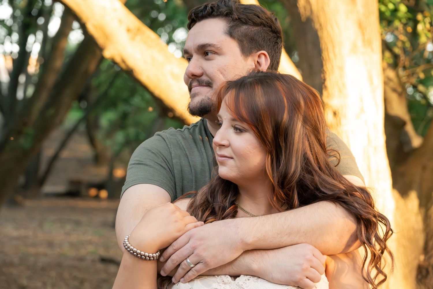 Couple embracing outdoors among trees in golden sunlight, man wrapping arms around woman with long auburn hair.