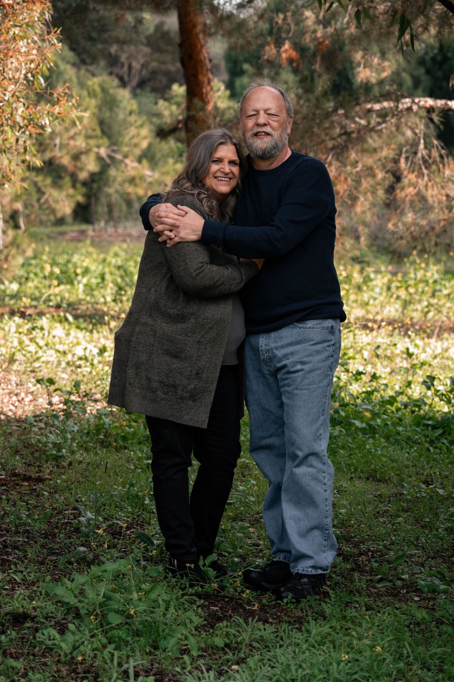 Full body portrait of an older couple hugging and smiling in a wooded park, standing on grass surrounded by trees during a relaxed outdoor