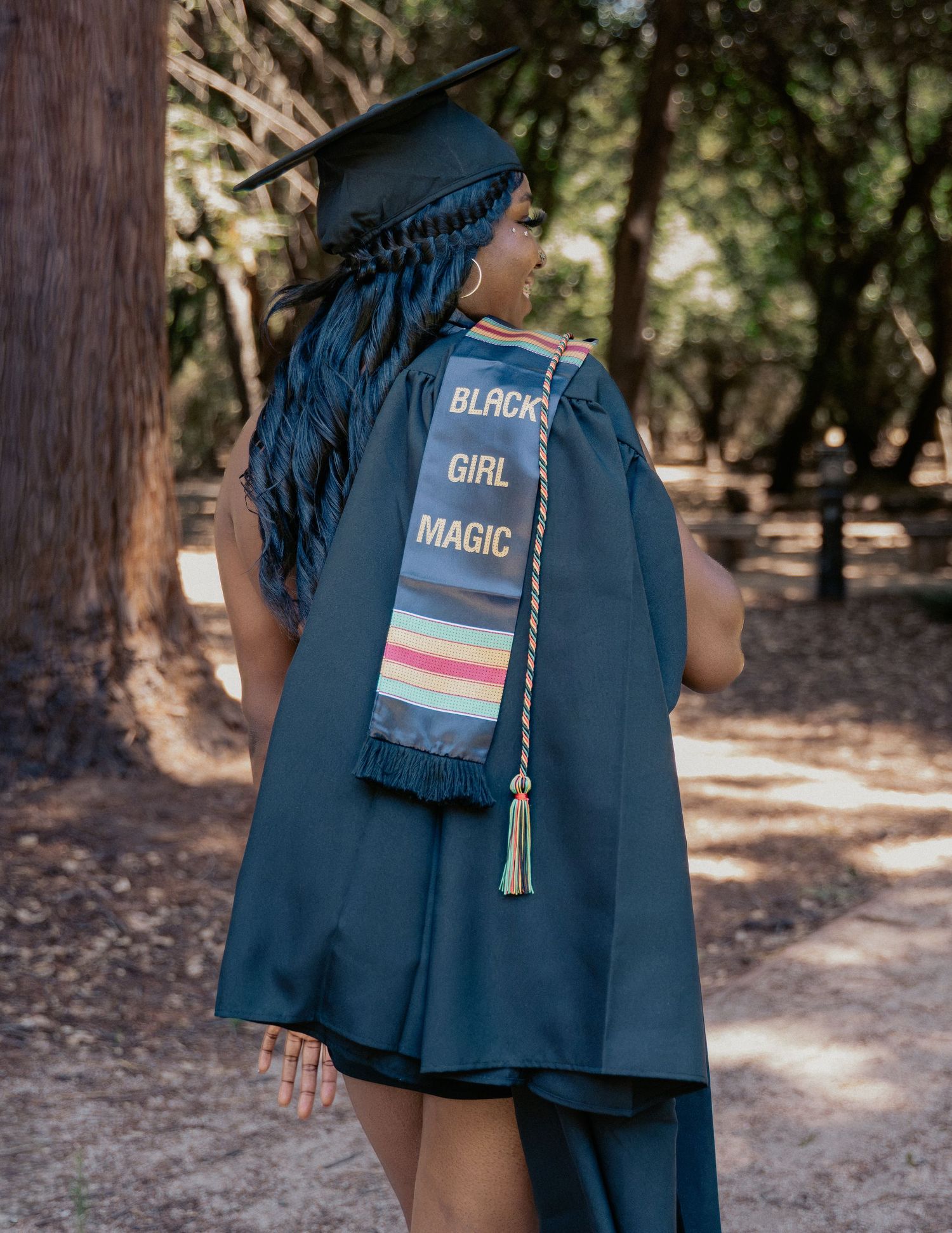 Graduate in navy cap and gown wearing a colorful Black Girl Magic stole, posing outdoors among trees.