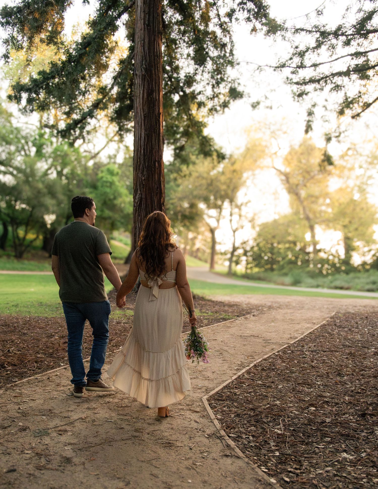 Couple holding hands walking along a park path surrounded by tall trees during golden hour, woman in white dress holding flowers.