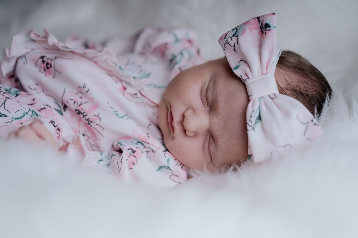Sleeping newborn baby in pink floral outfit with matching bow headband on white fluffy blanket.
