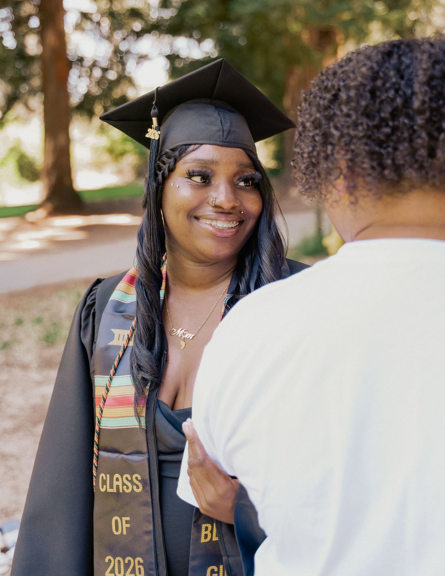 A smiling graduate in cap and gown with Class of 2026 stole talks with another person outdoors.