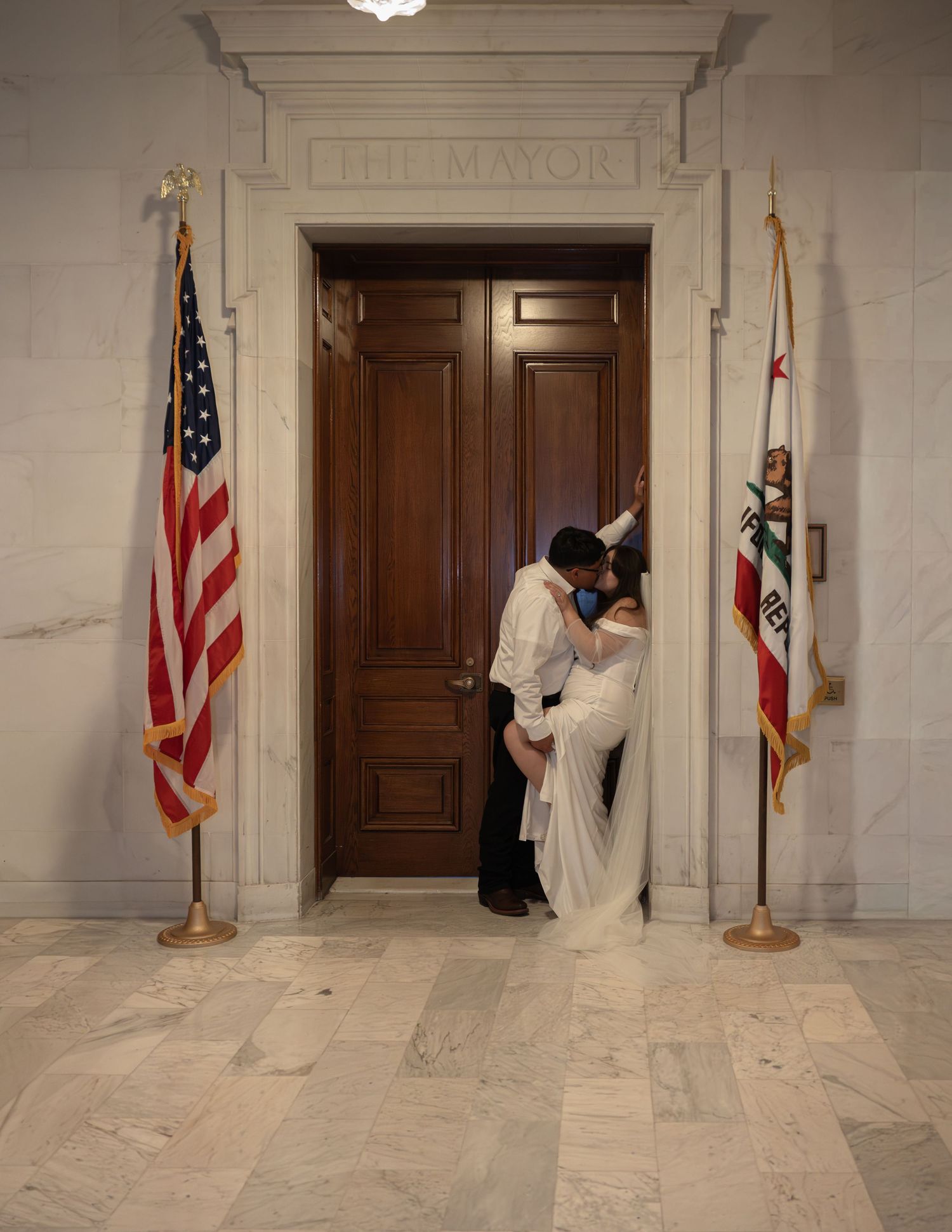 Couple in wedding attire kissing outside The Mayor's office, flanked by US and California state flags. Elopemen photography in San Francisco