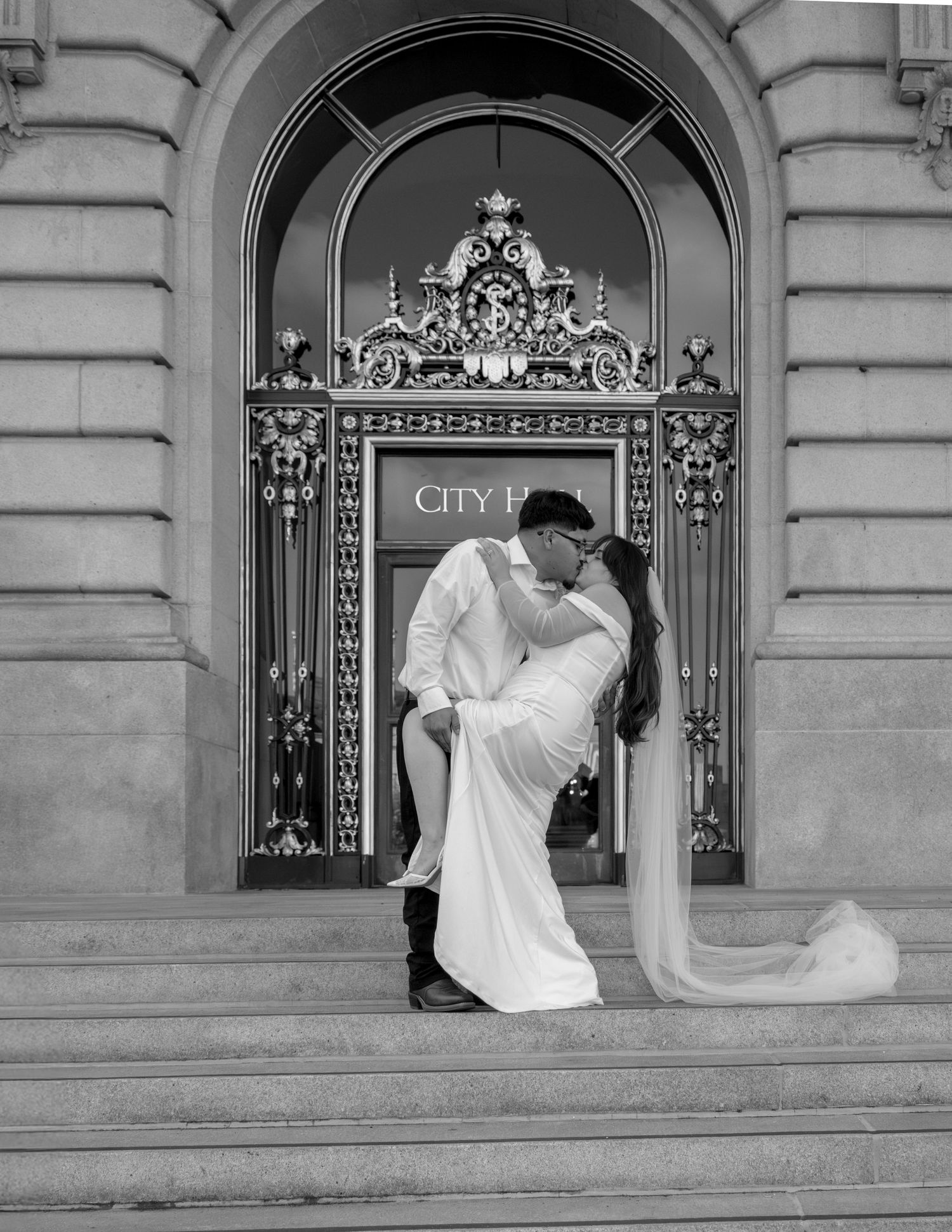 Black and white photo of a couple sharing a romantic dip kiss on the steps of City Hall in wedding attire.