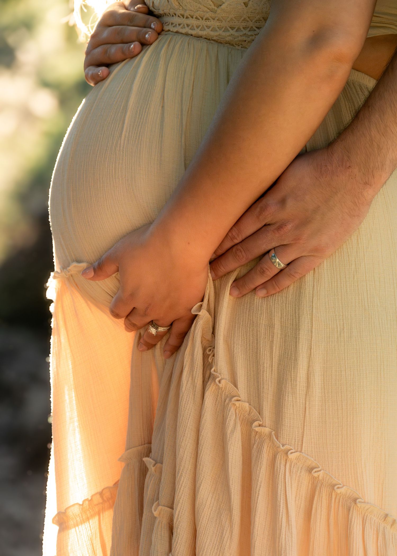 Pregnant woman in flowy cream dress holding hands with partner, showcasing baby bump and engagement ring in warm golden light. Bump-to-baby.