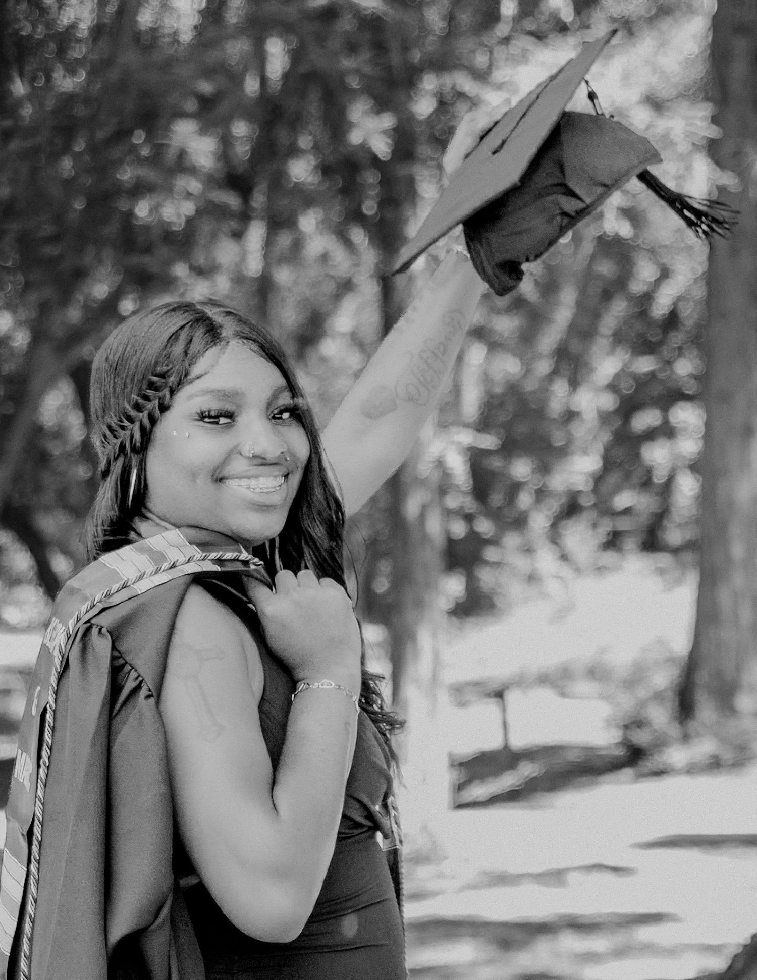 Black and white photo of a smiling graduate holding her cap in the air outdoors among trees.