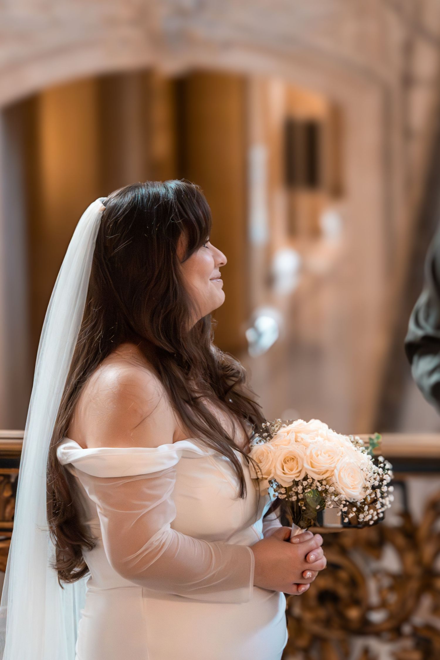 A bride in an off-shoulder wedding dress holds a peach bouquet in an elegant venue.