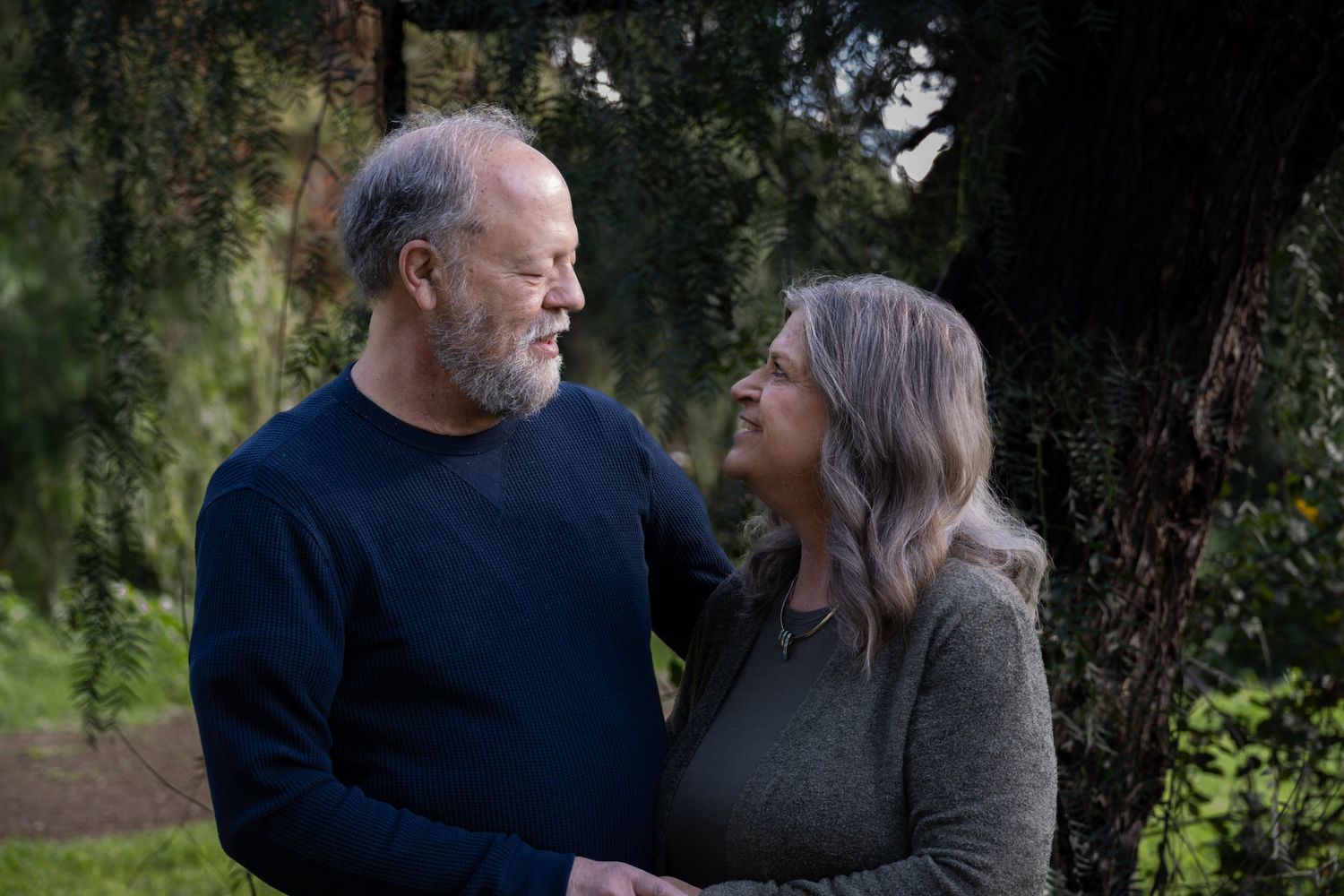 An older couple stands together outdoors near a tree, smiling warmly at each other in a serene garden setting.