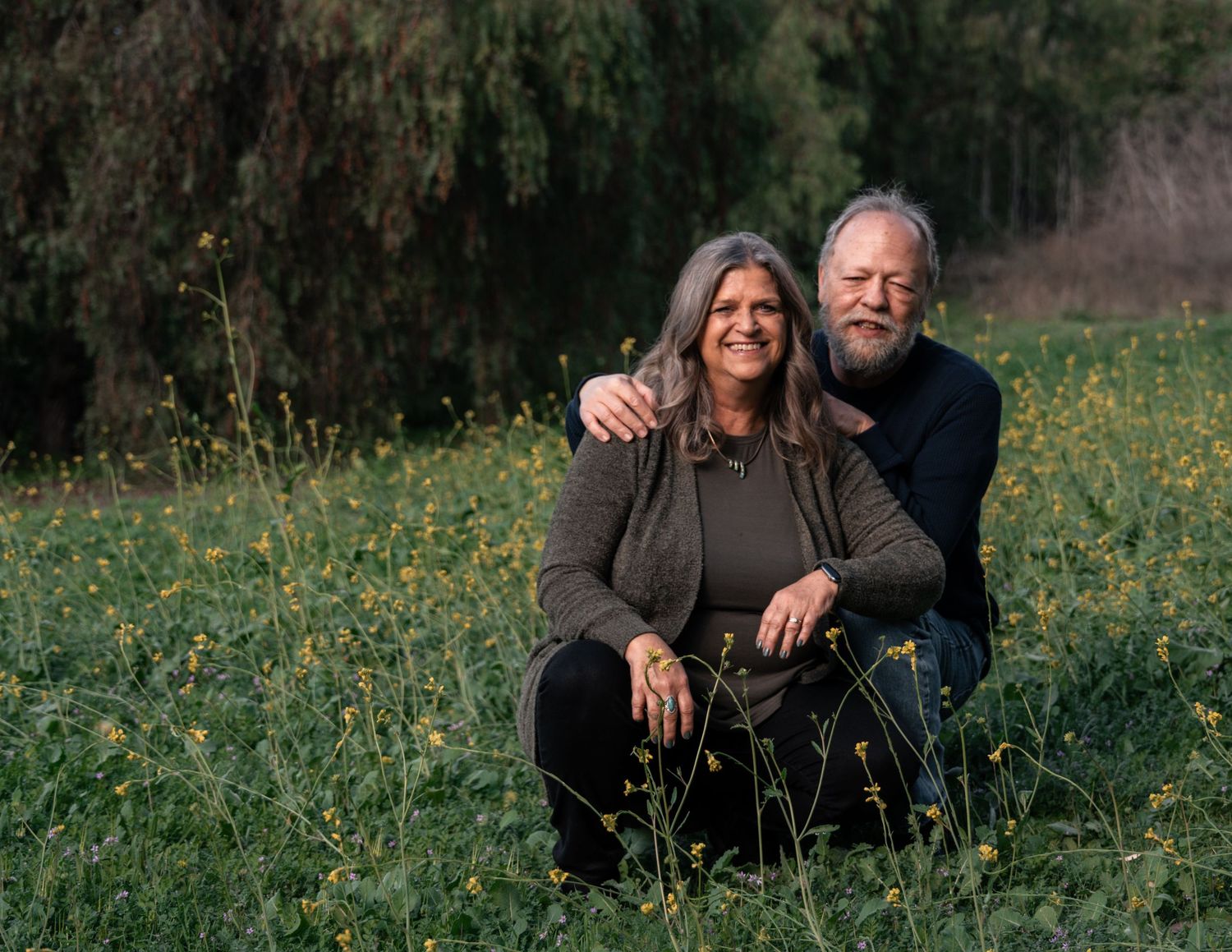 A couple sits together in a field of yellow wildflowers surrounded by trees.