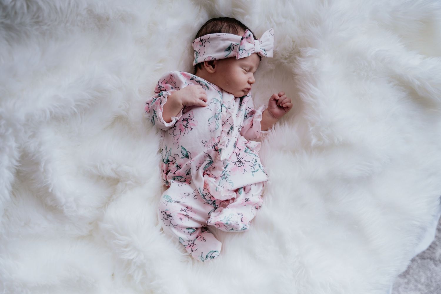 Newborn baby girl sleeping on white fluffy blanket wearing pink floral romper and matching bow headband. Newborn portraits, in-home session.