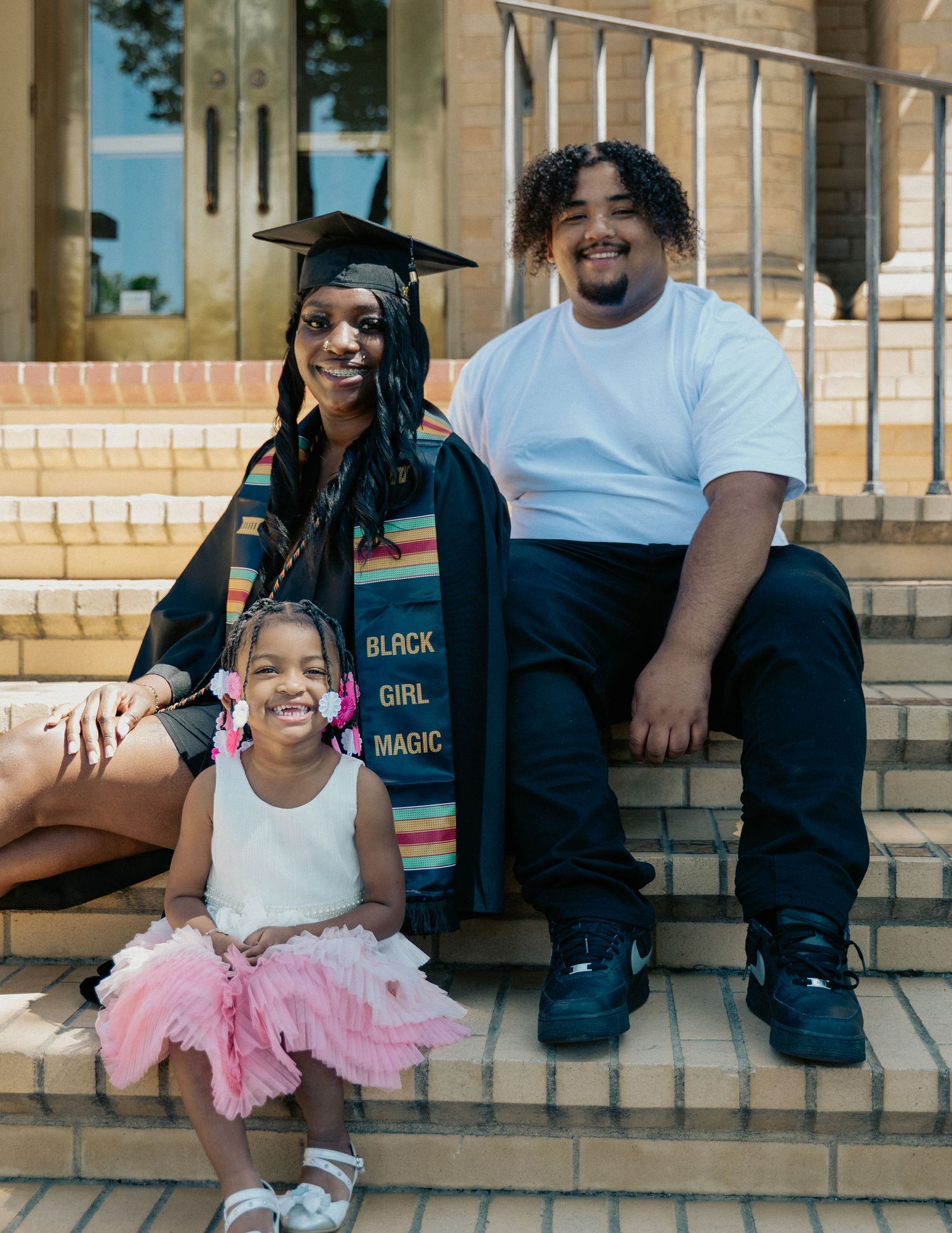 Graduate in cap and gown with Black Girl Magic stole poses on steps with young girl in tutu and young man.