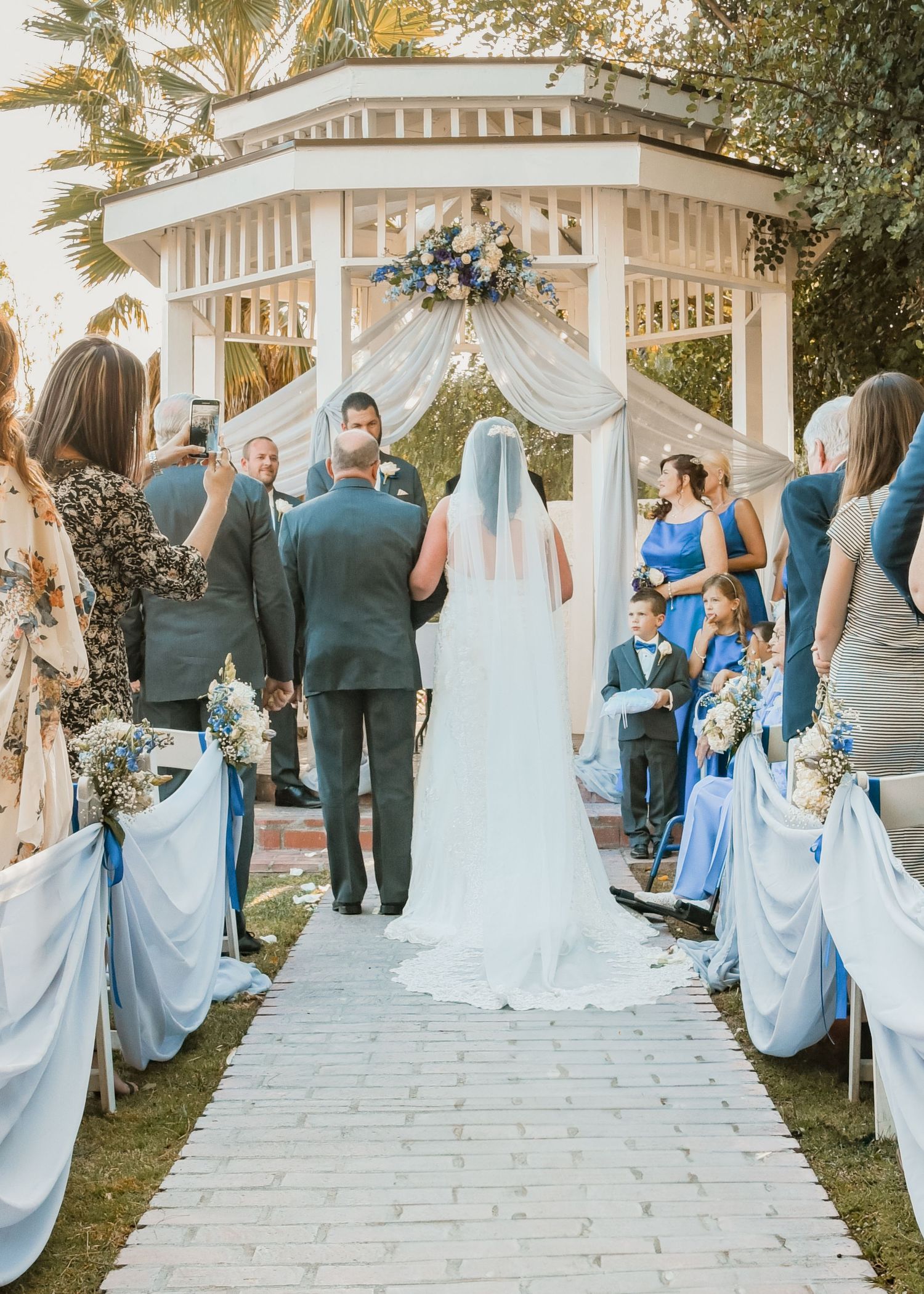 Outdoor wedding ceremony at a white gazebo with guests in blue dresses seated on white chairs.