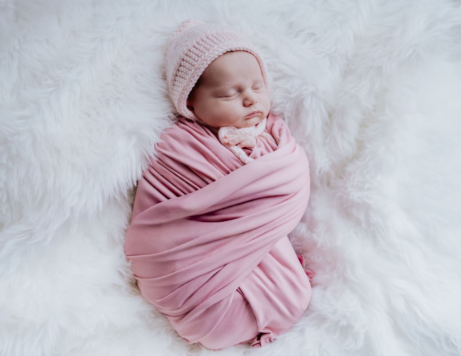 Newborn baby girl swaddled in pink wrap and knit bonnet, sleeping peacefully on white fluffy blanket.