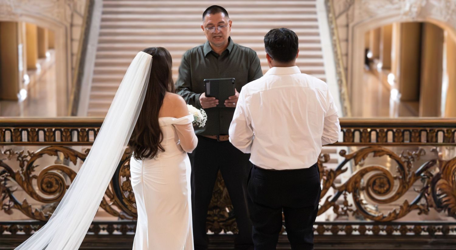 Couple exchanging vows with officiant on ornate staircase during intimate wedding ceremony at San Francisco City Hall in San Francisco, CA.