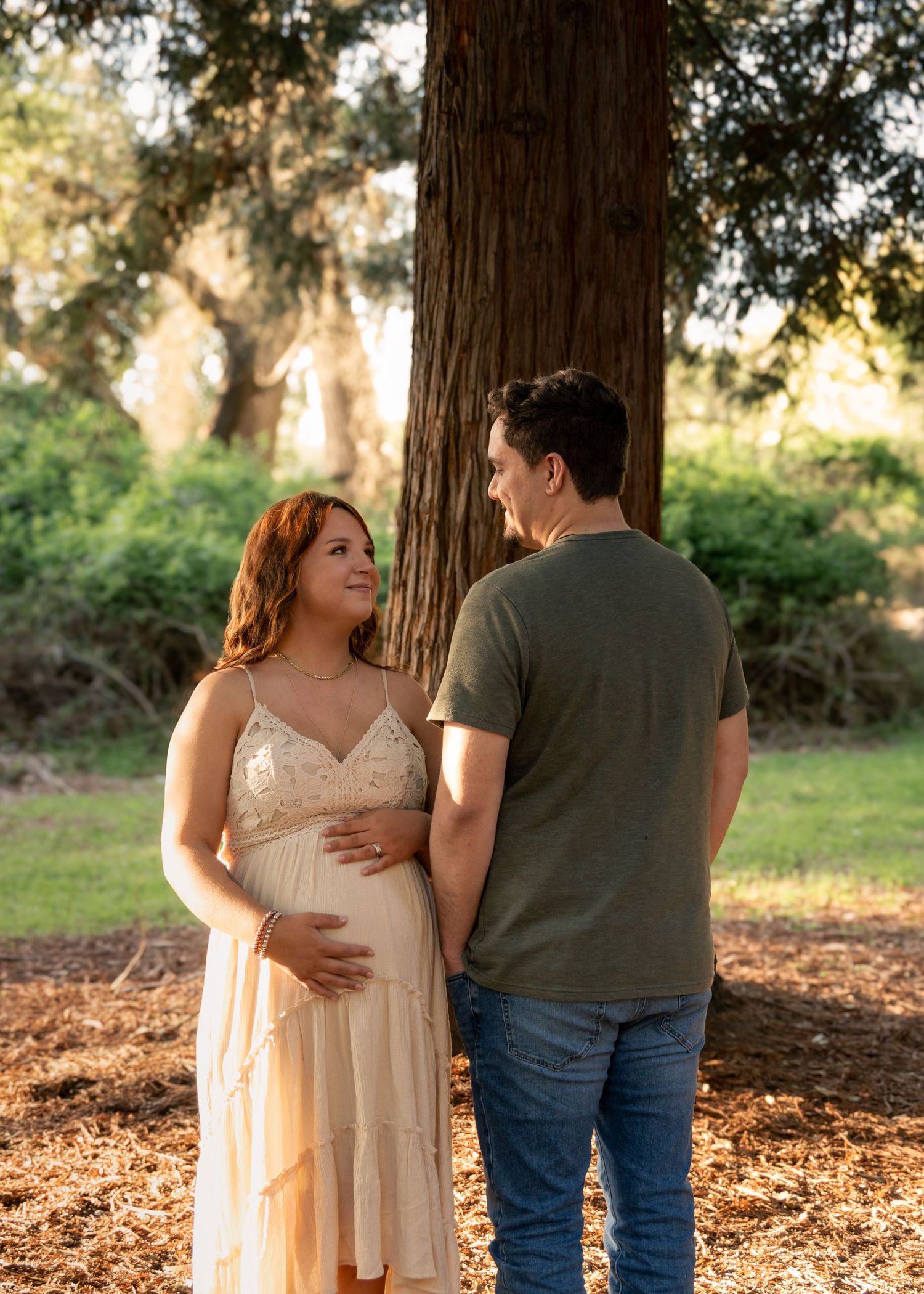 A couple in casual attire embraces while standing among fallen leaves beneath tall trees in a natural outdoor setting.