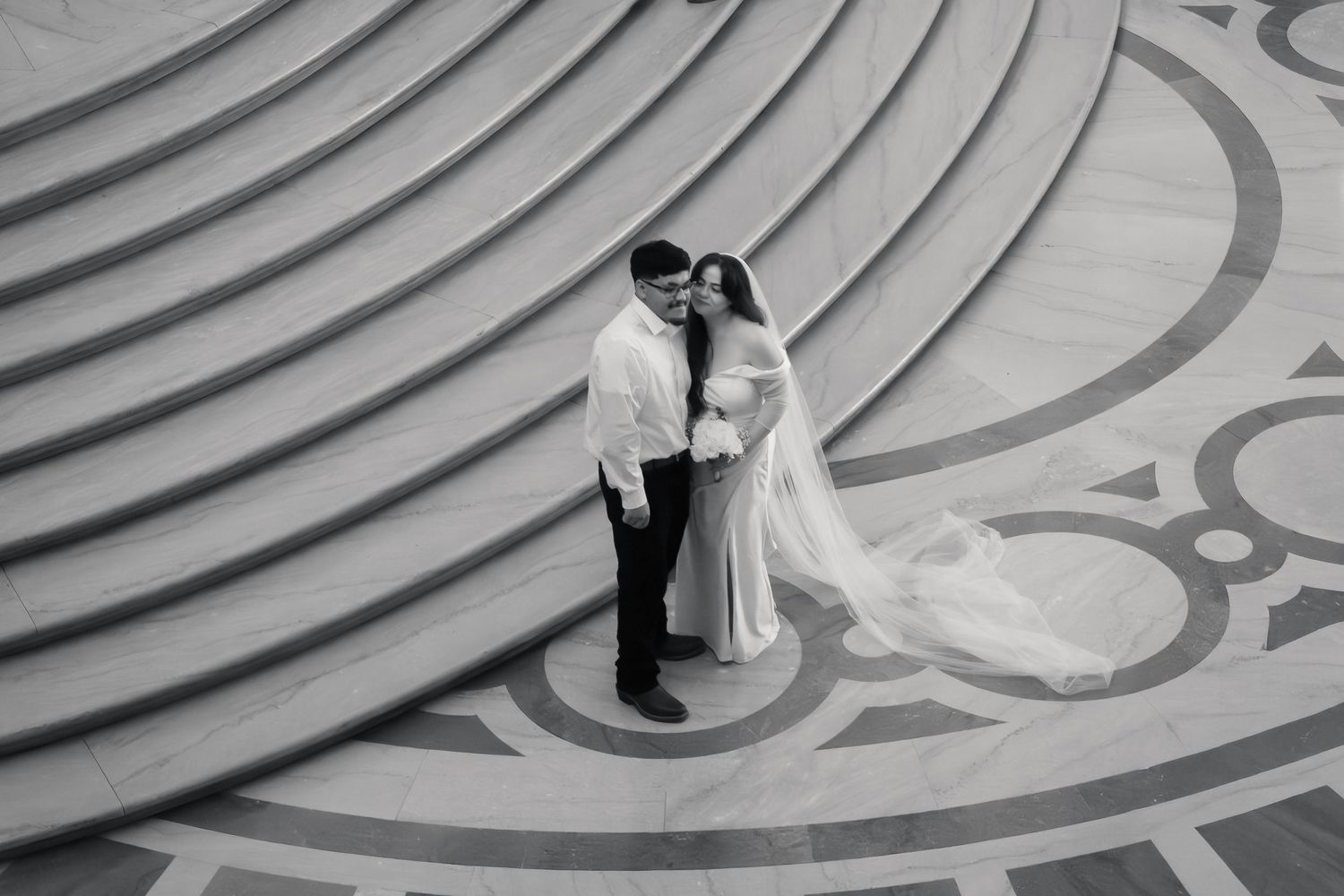 Black and white aerial photo of bride and groom embracing on ornate circular patterned floor with curved steps.