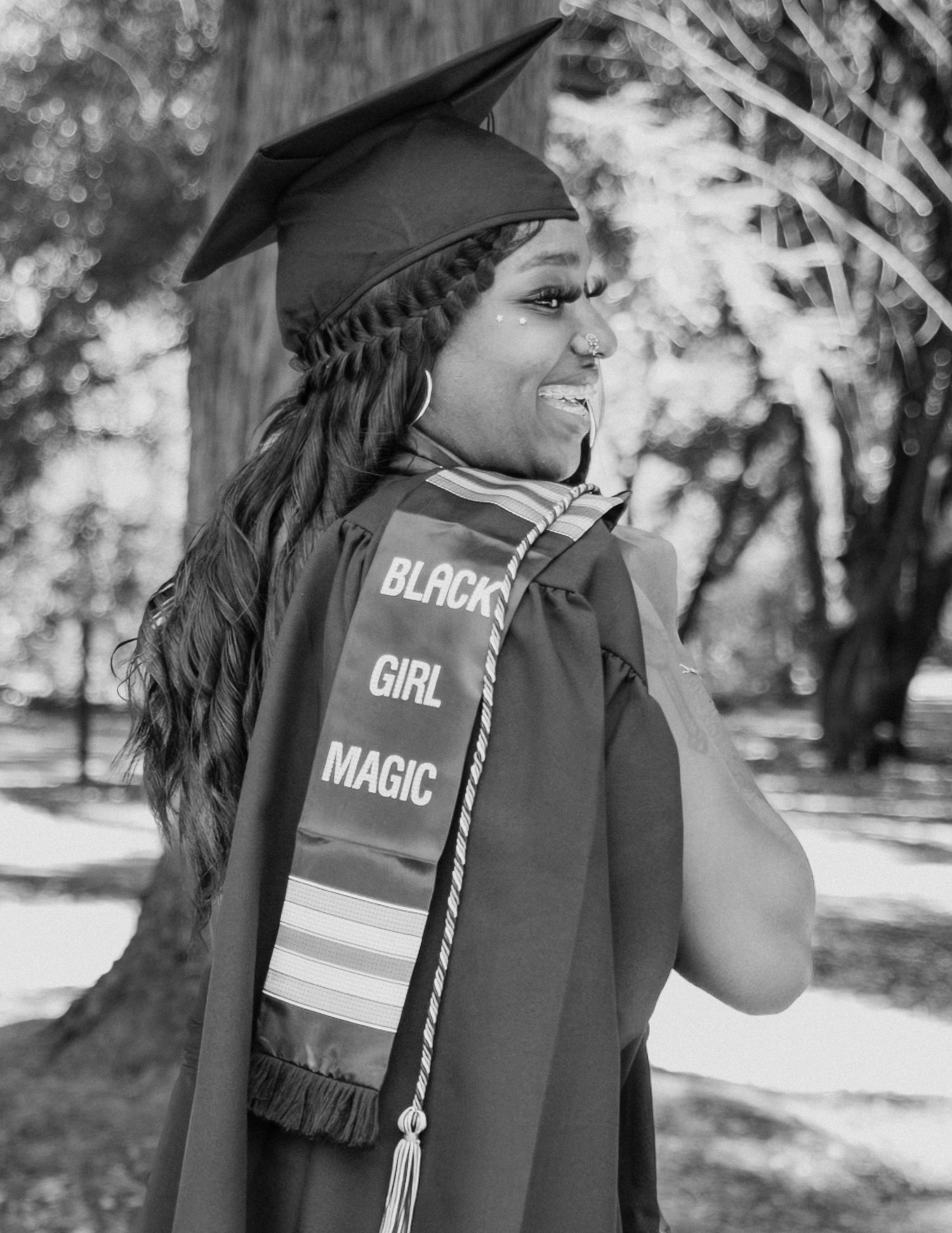 Black woman in graduation cap and gown with Black Girl Magic stole smiles outdoors in black and white photo.