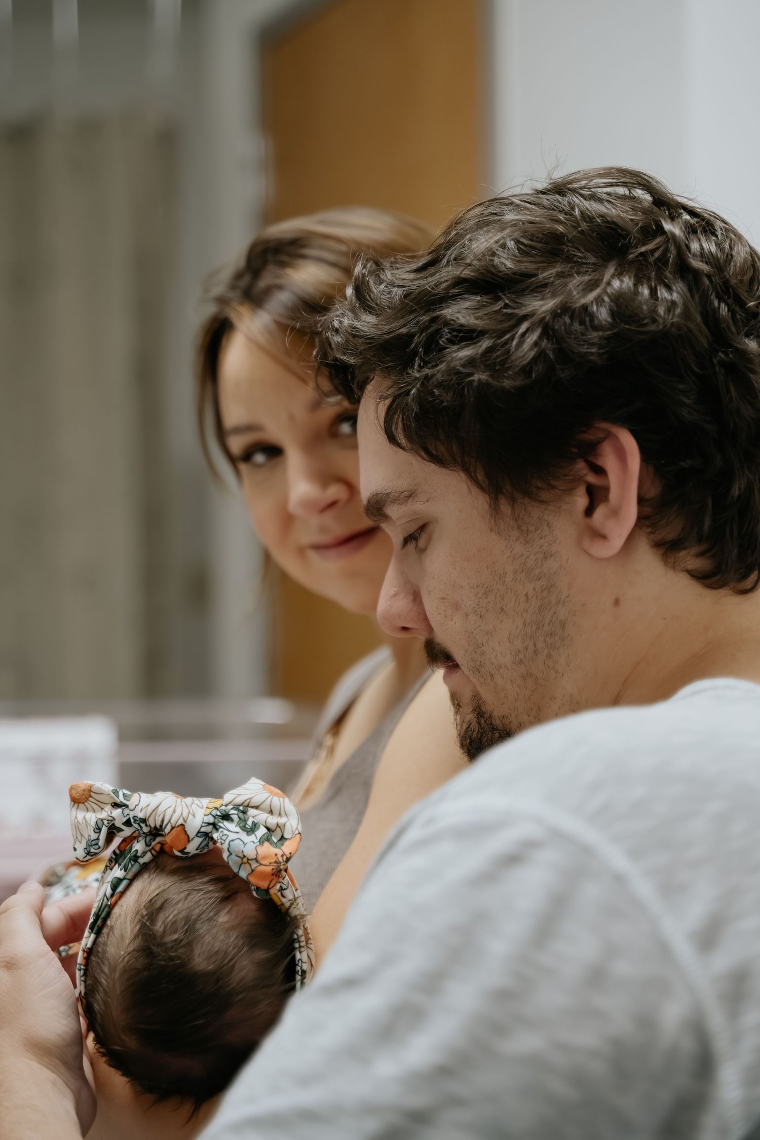 A young couple lovingly gazes at their newborn baby wearing a floral headband in a hospital room.