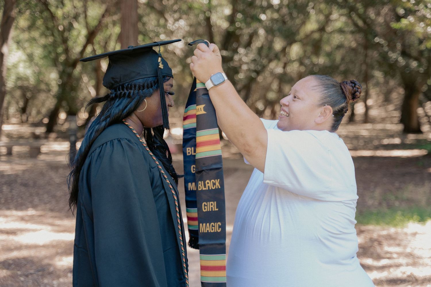 Graduate in cap and gown receives stole from woman outdoors among trees at graduation celebration.