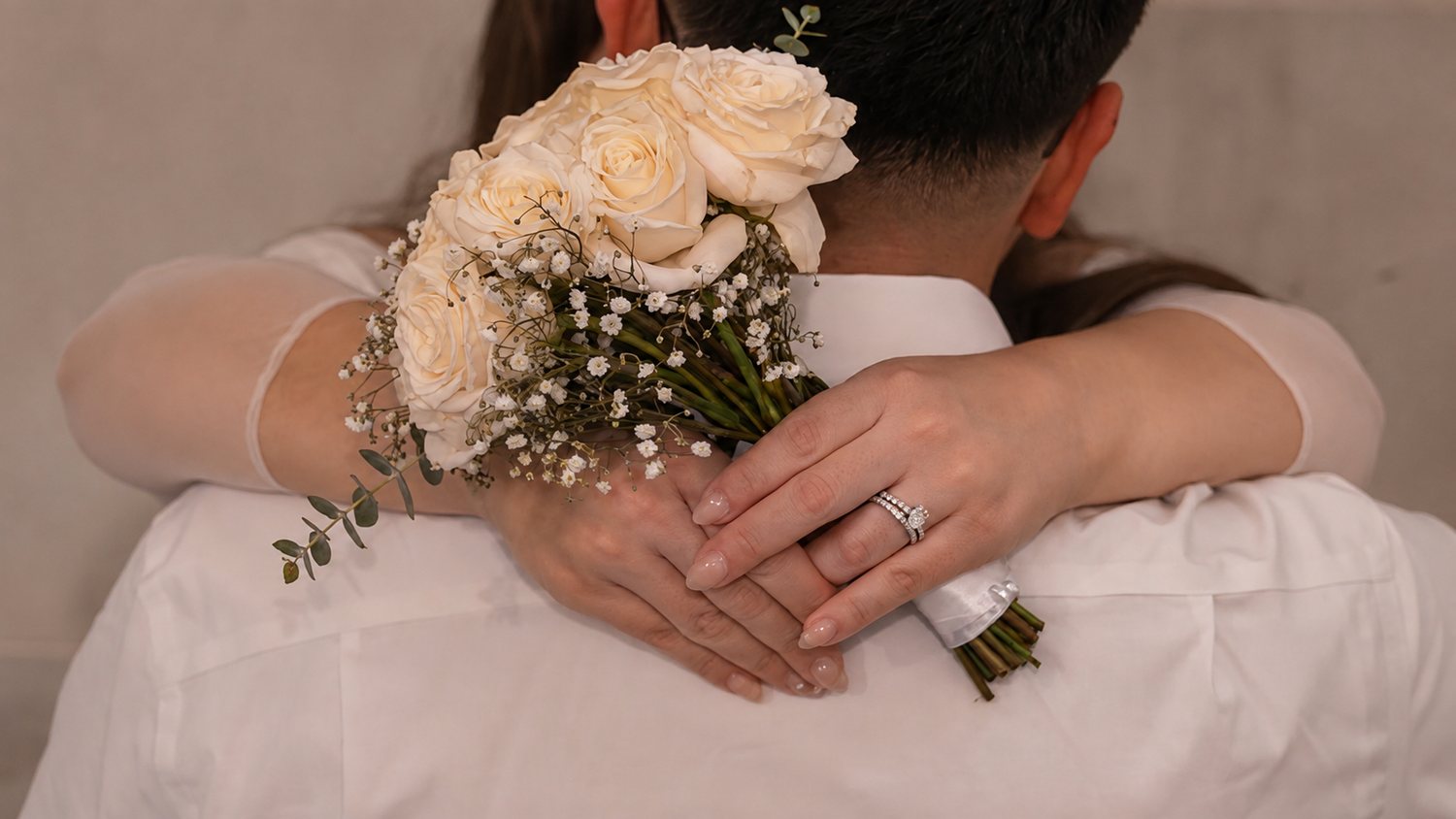 Bride holding white rose bouquet while embracing groom, wedding ring visible on her finger.