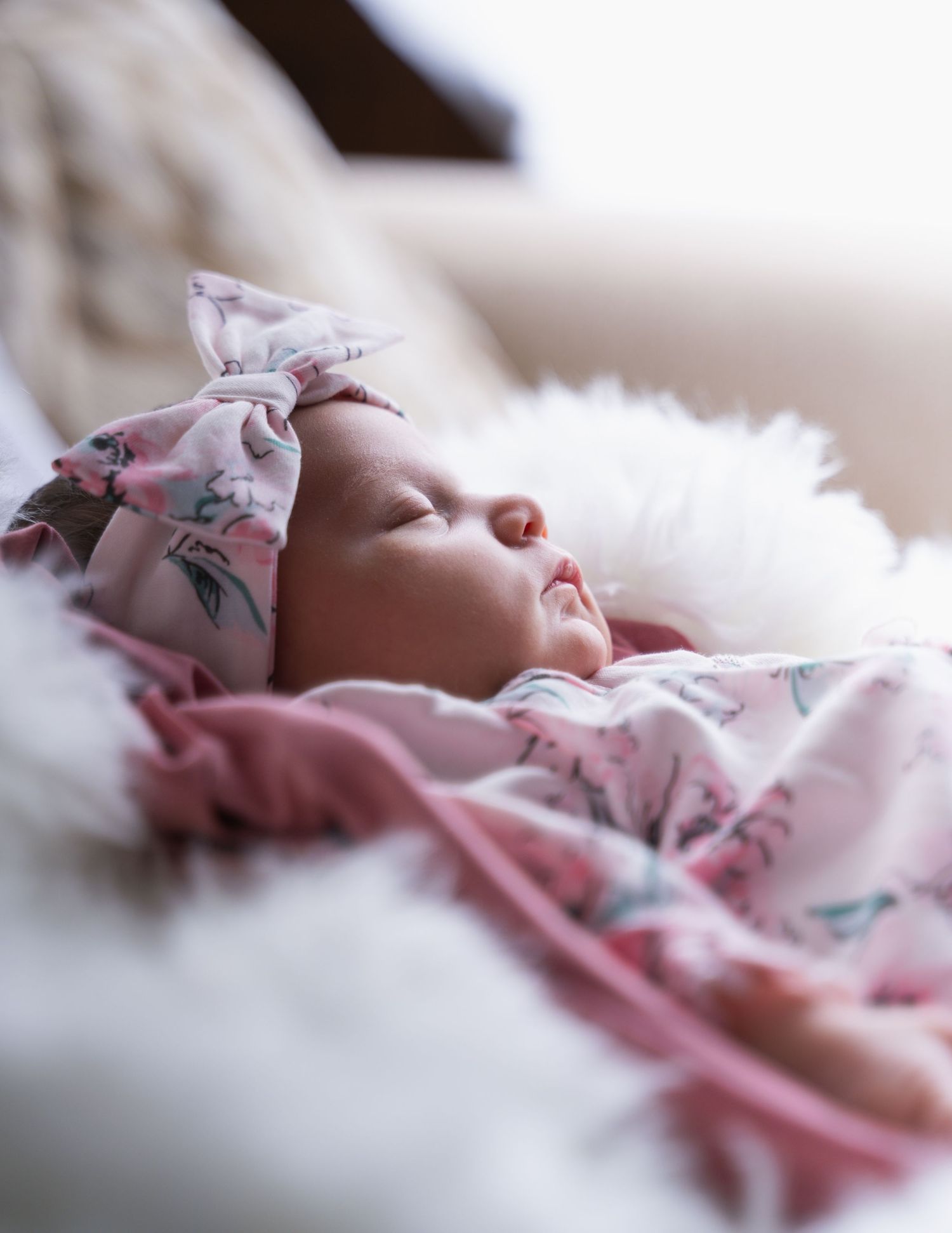 Sleeping newborn baby girl in pink floral swaddle blanket and matching bow headband on white fluffy surface.