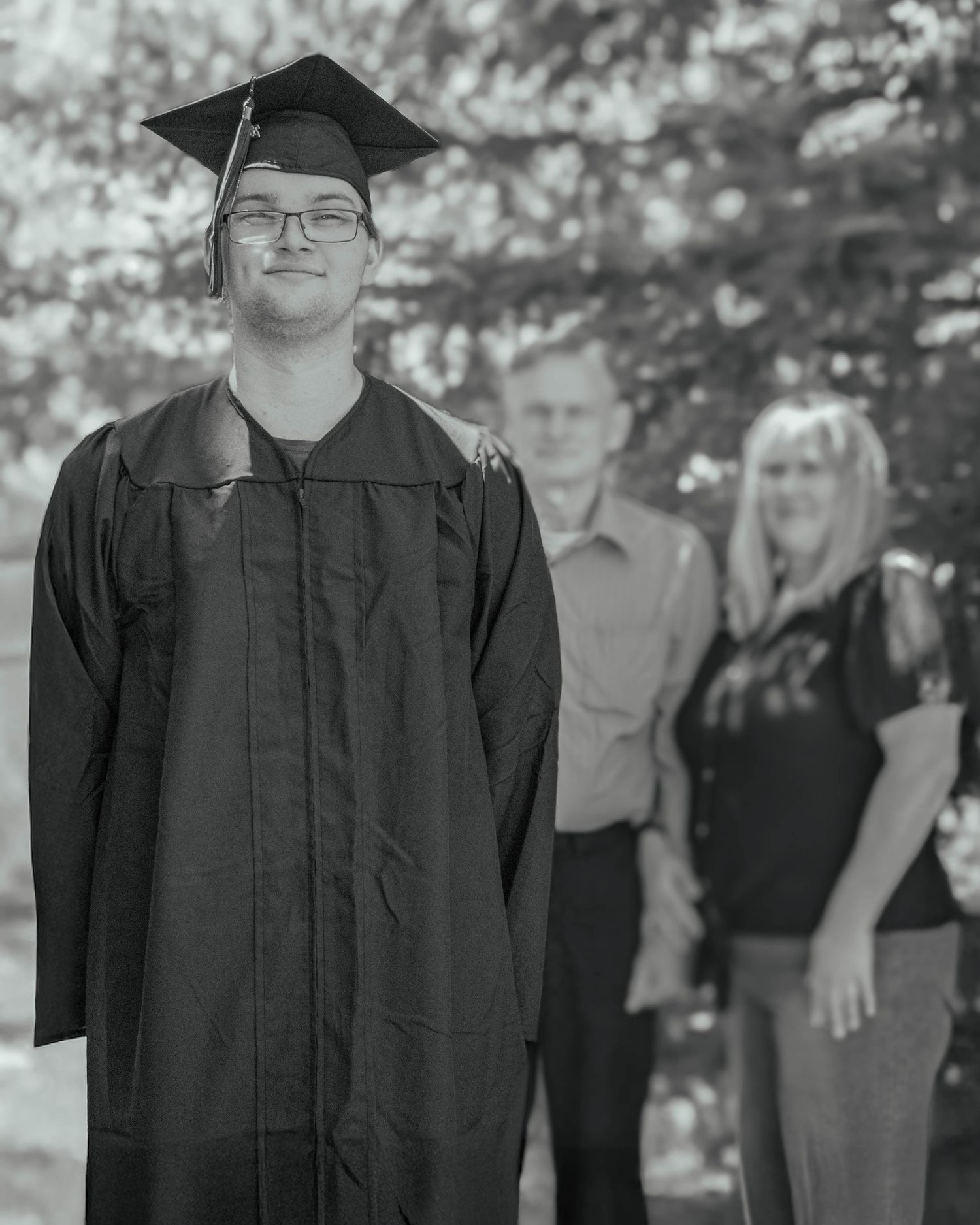 A young graduate in cap and gown smiles proudly outdoors, with two adults standing proudly behind him.