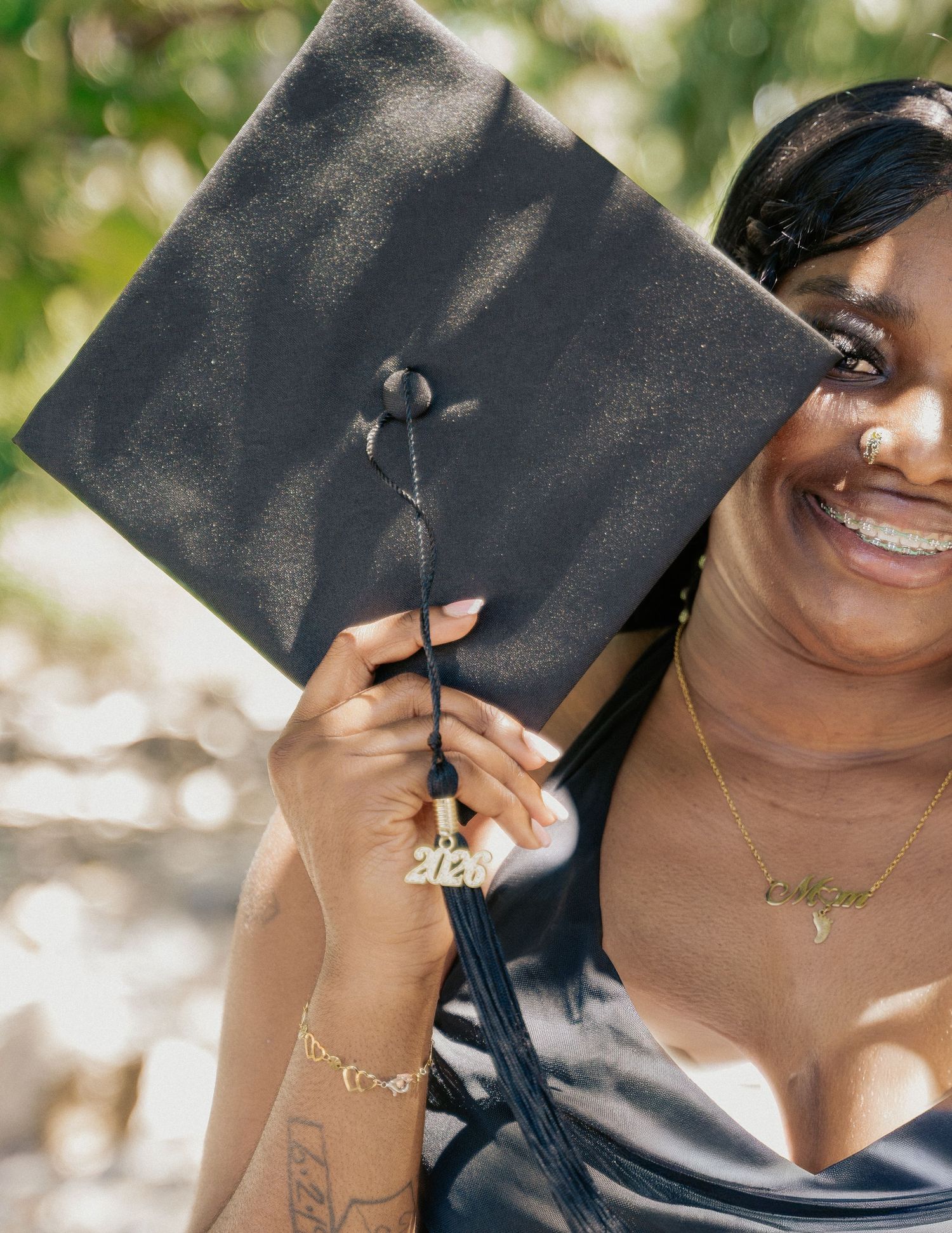 Smiling graduate holding black mortarboard cap with 2020 tassel, wearing gold jewelry and blue dress outdoors.