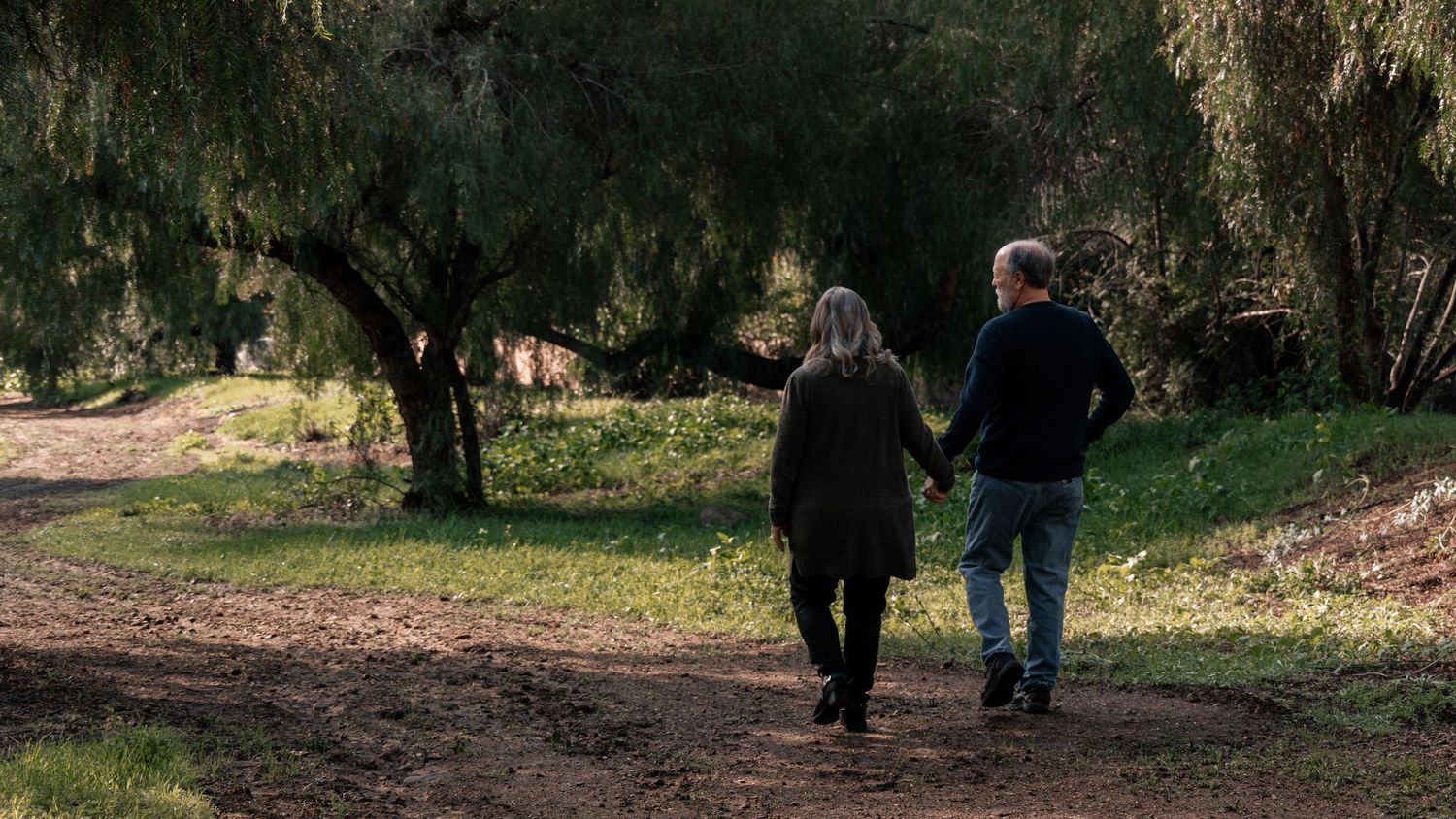 Elderly couple holding hands walking on a dirt path through a serene wooded park area.