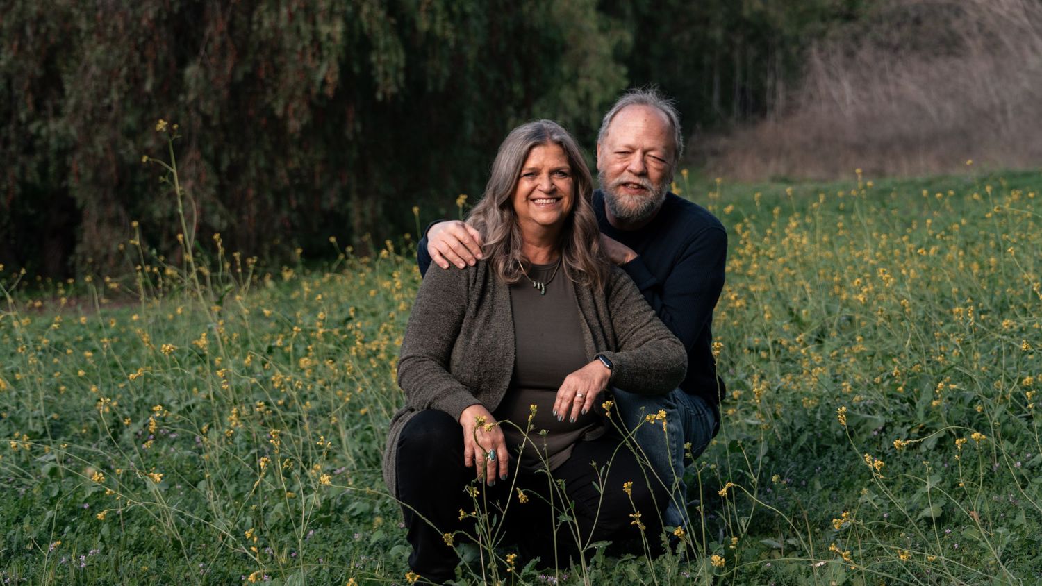 A smiling couple sits together in a lush green meadow with wildflowers, sharing a warm embrace outdoors. Dixon, CA