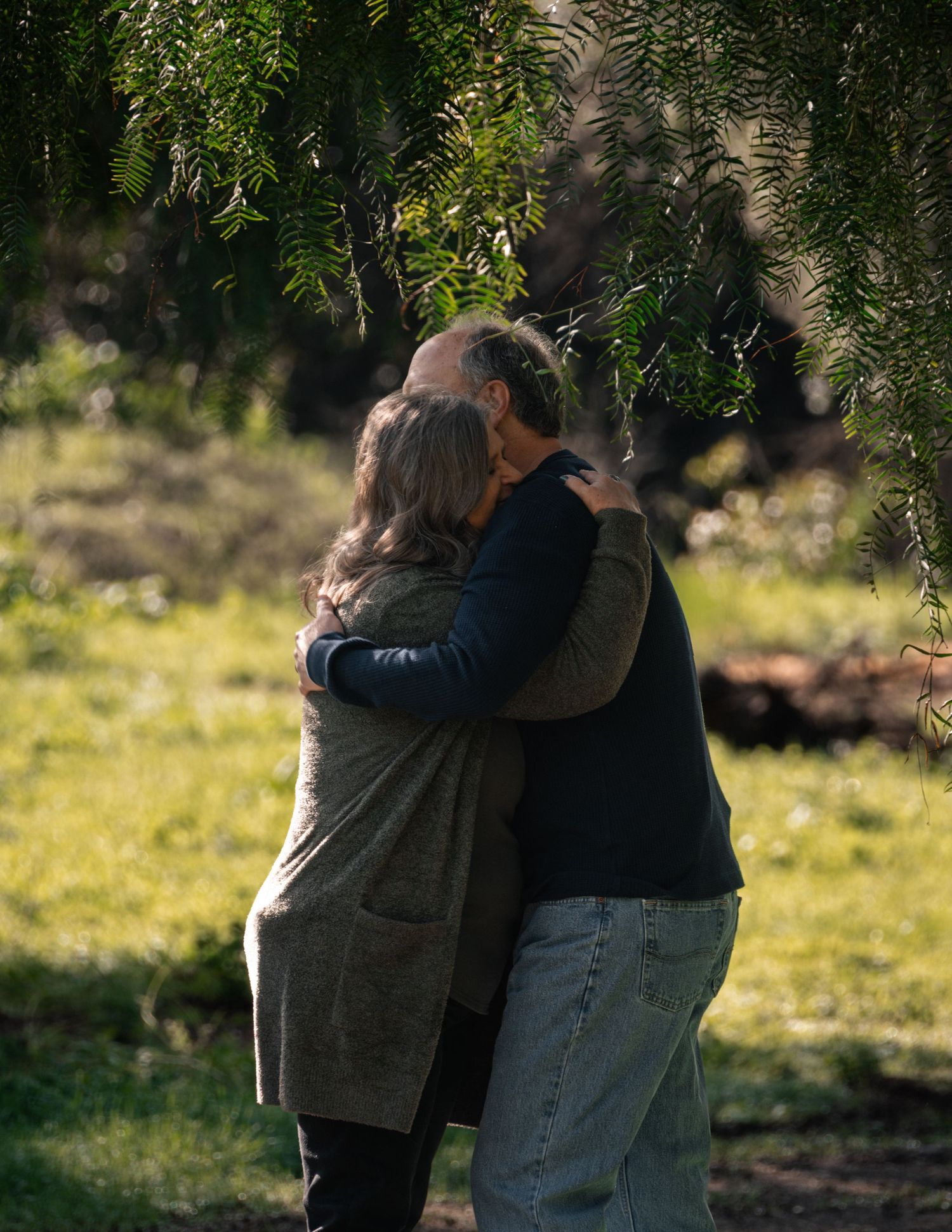 A couple embraces tenderly in a sunlit outdoor setting with trees and grass in the background.
