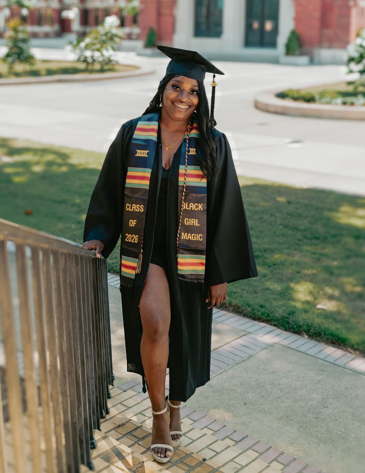 A smiling graduate in black cap and gown with colorful kente stole reading Class of 2025 and Black Girl Magic poses outdoors.