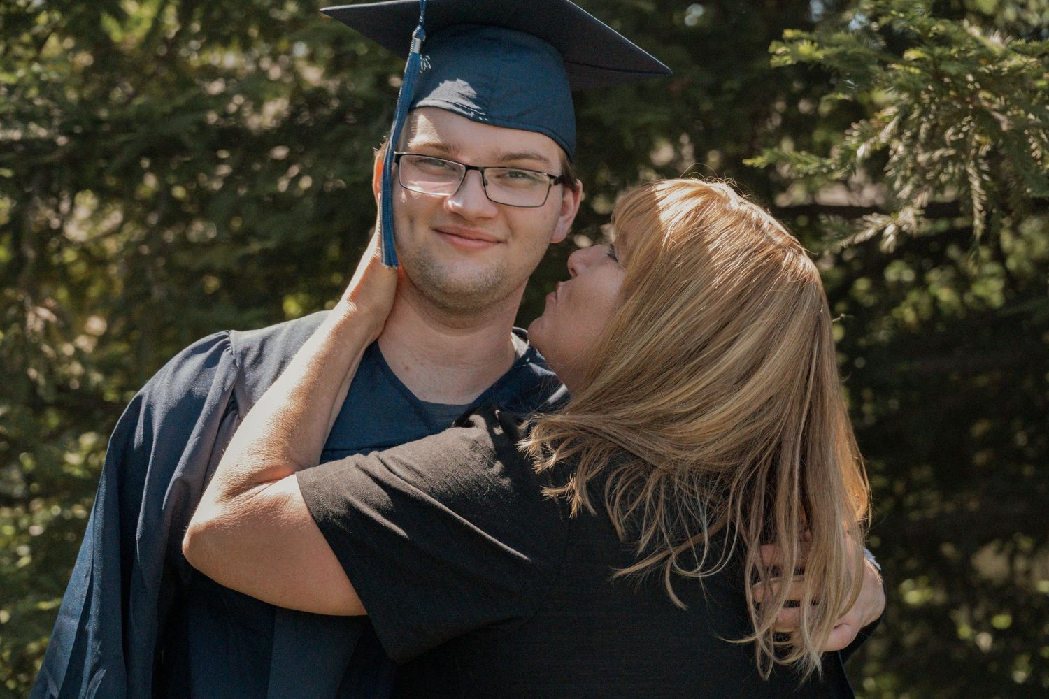 Graduate in cap and gown smiling as mother kisses his cheek, outdoor senior photos near Roseville CA by Jolie & Rae Photography