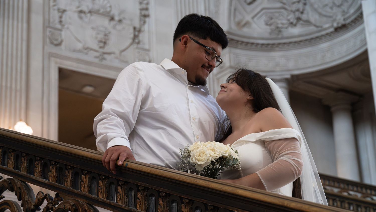 Newlywed couple in wedding attire sharing a romantic moment on a balcony at San Francisco City Hall.